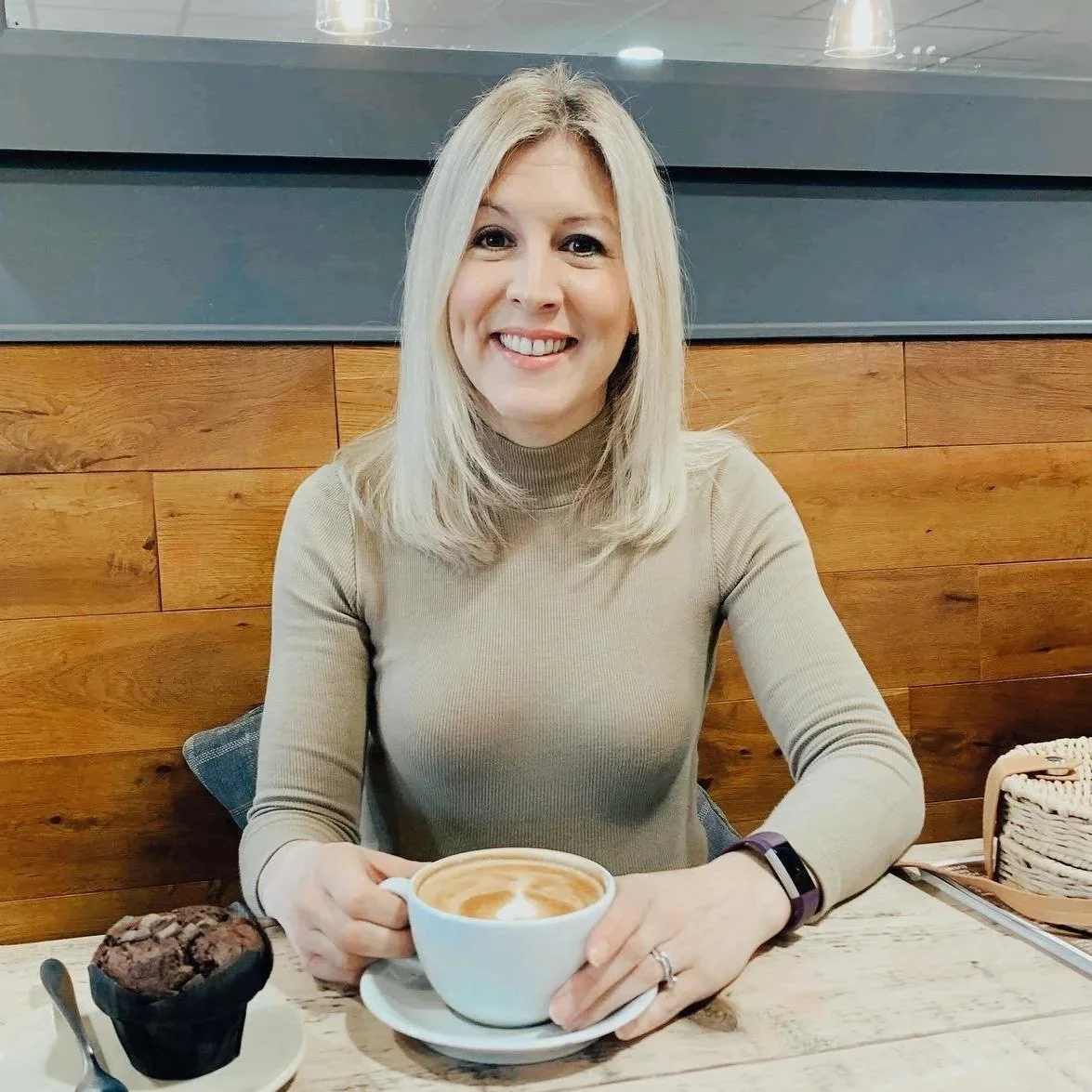 A woman with blonde hair sitting at a table in a cafe, smiling, holding a cup of coffee with latte art, next to a chocolate muffin.