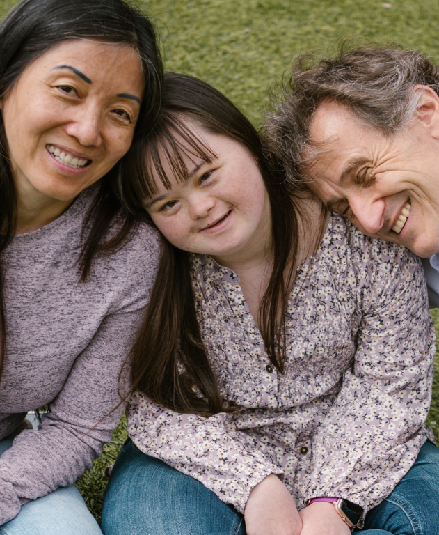 A diverse family of three sitting on the grass and smiling at the camera.