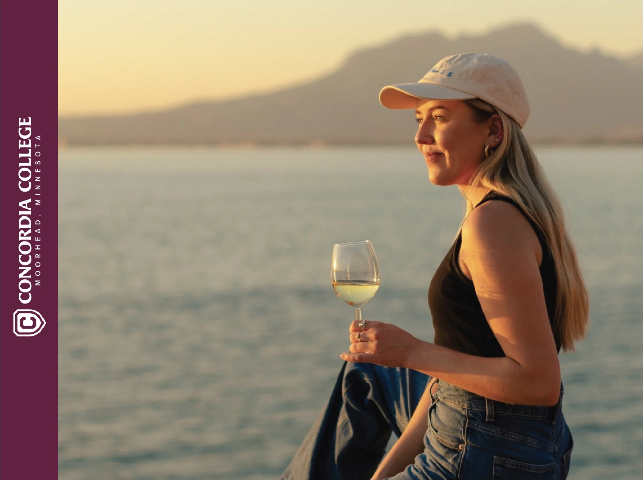 A woman sitting by a lake during sunset, holding a glass of white wine, wearing a beige cap, black tank top, and jeans.