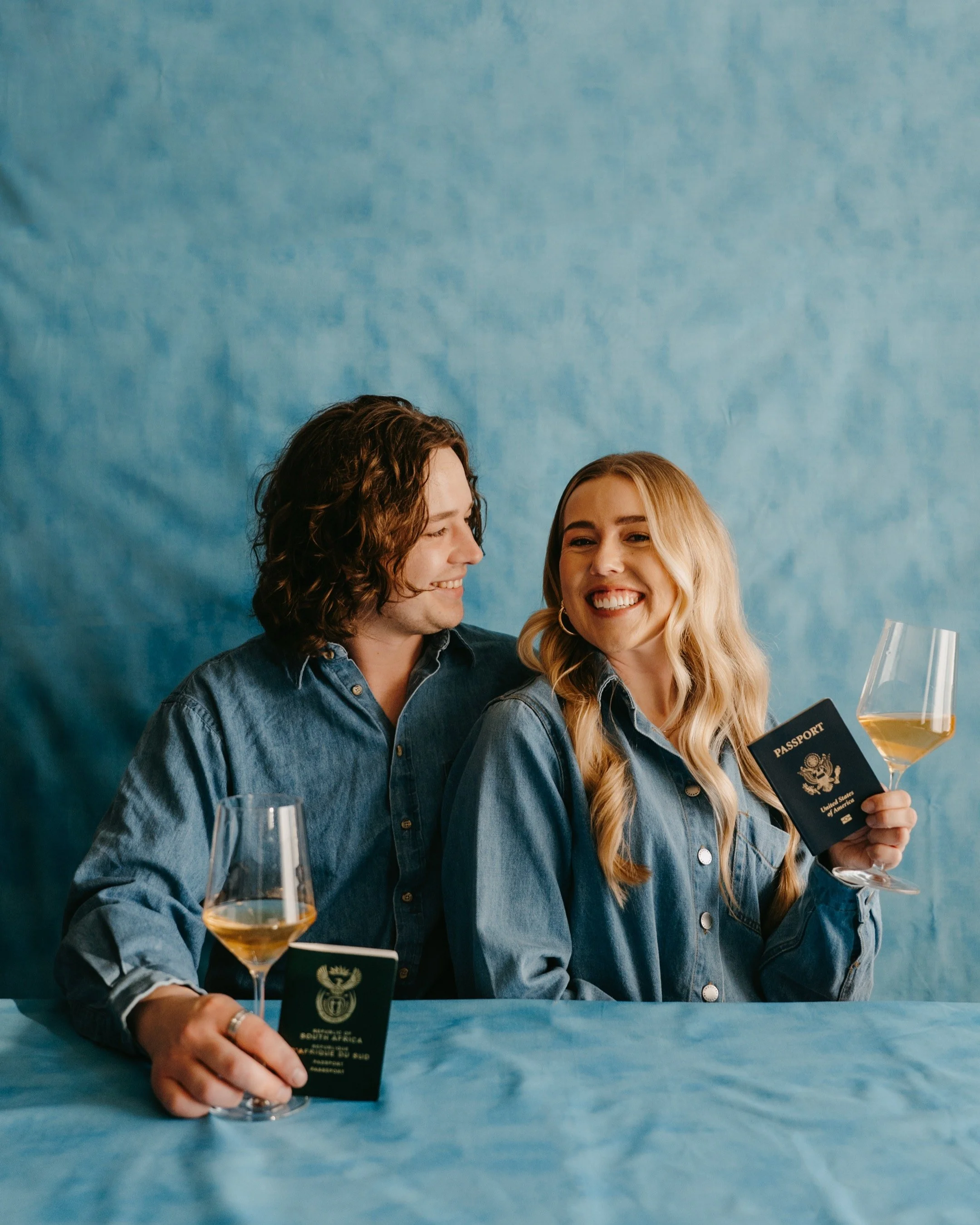 A smiling couple holding wine glasses and passports, sitting together against a blue textured background.