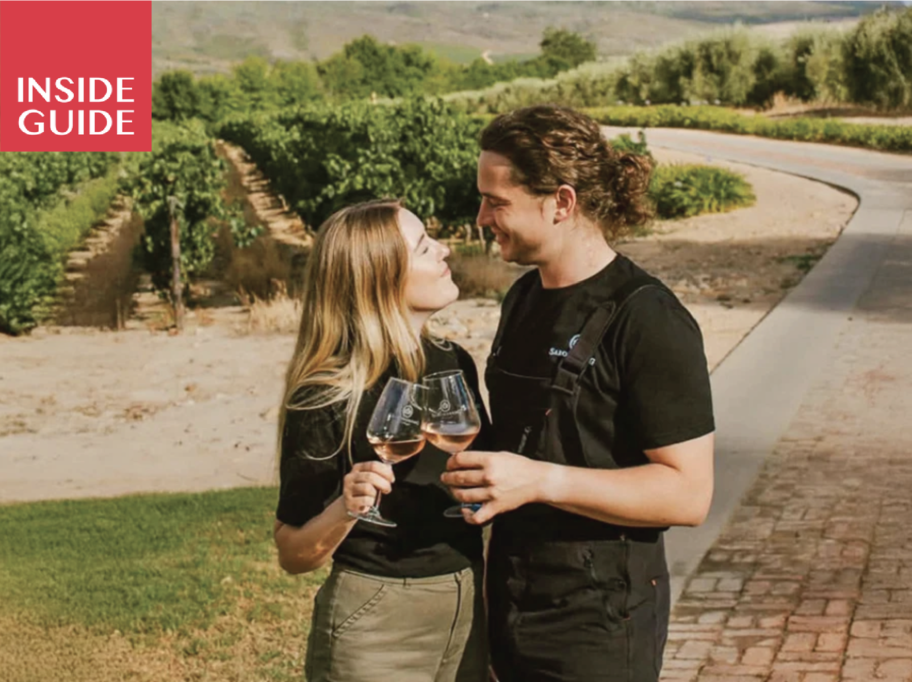 A young couple standing outdoors in a vineyard holding glasses of rosé wine, smiling at each other with lush green rows of grapevines in the background.