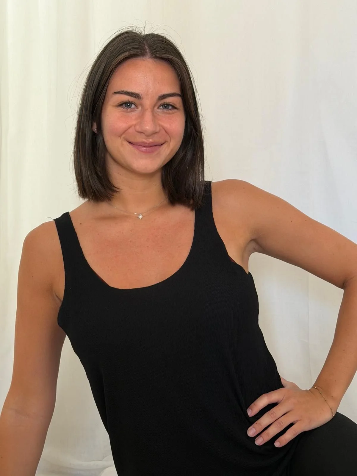 Jeune femme aux cheveux bruns, portant un top noir, souriant, avec une main sur la hanche, devant un fond blanc.