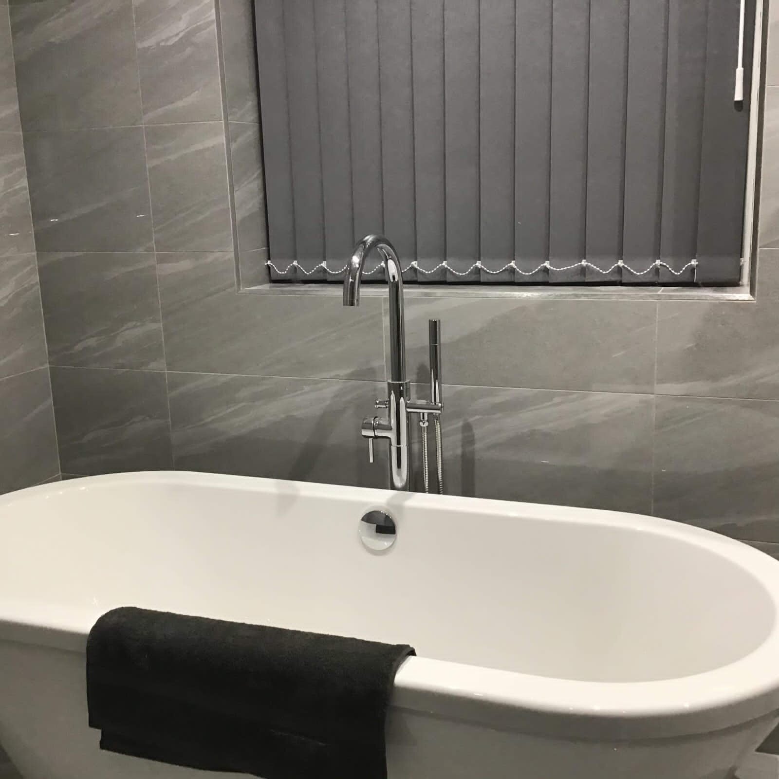 Modern bathroom with a white freestanding bathtub, a black towel draped over it, gray tiled walls, a window with closed gray blinds, and a chrome faucet with a hand shower.