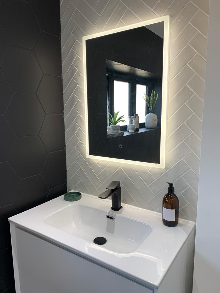 Bathroom with a white sink, black faucet, and brown soap dispenser. Above the sink is a rectangular mirror with LED lighting, reflected in a window with potted plants.