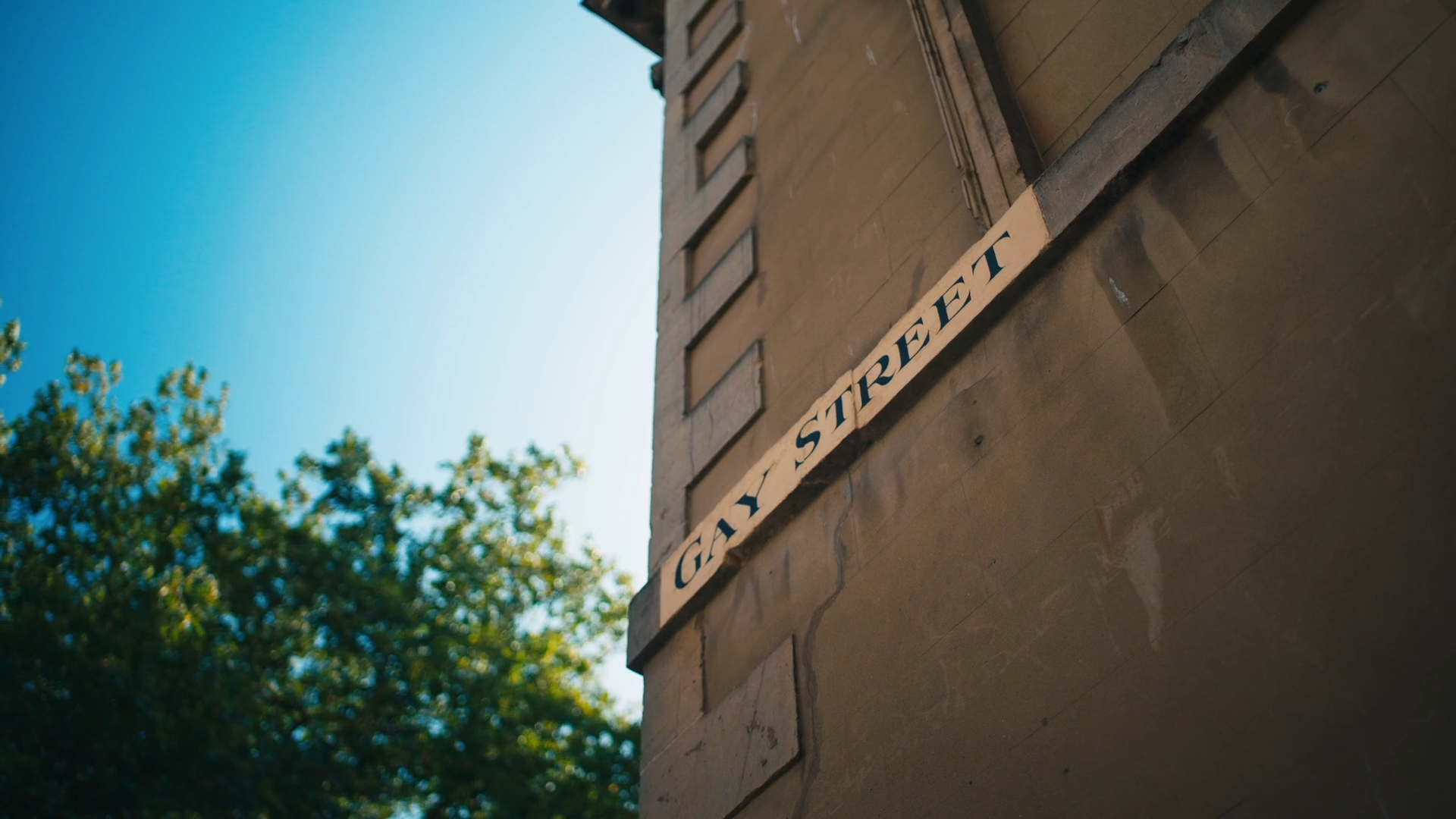 Street sign on a brick wall that reads 'GAY STREET' with trees and a clear blue sky in the background.