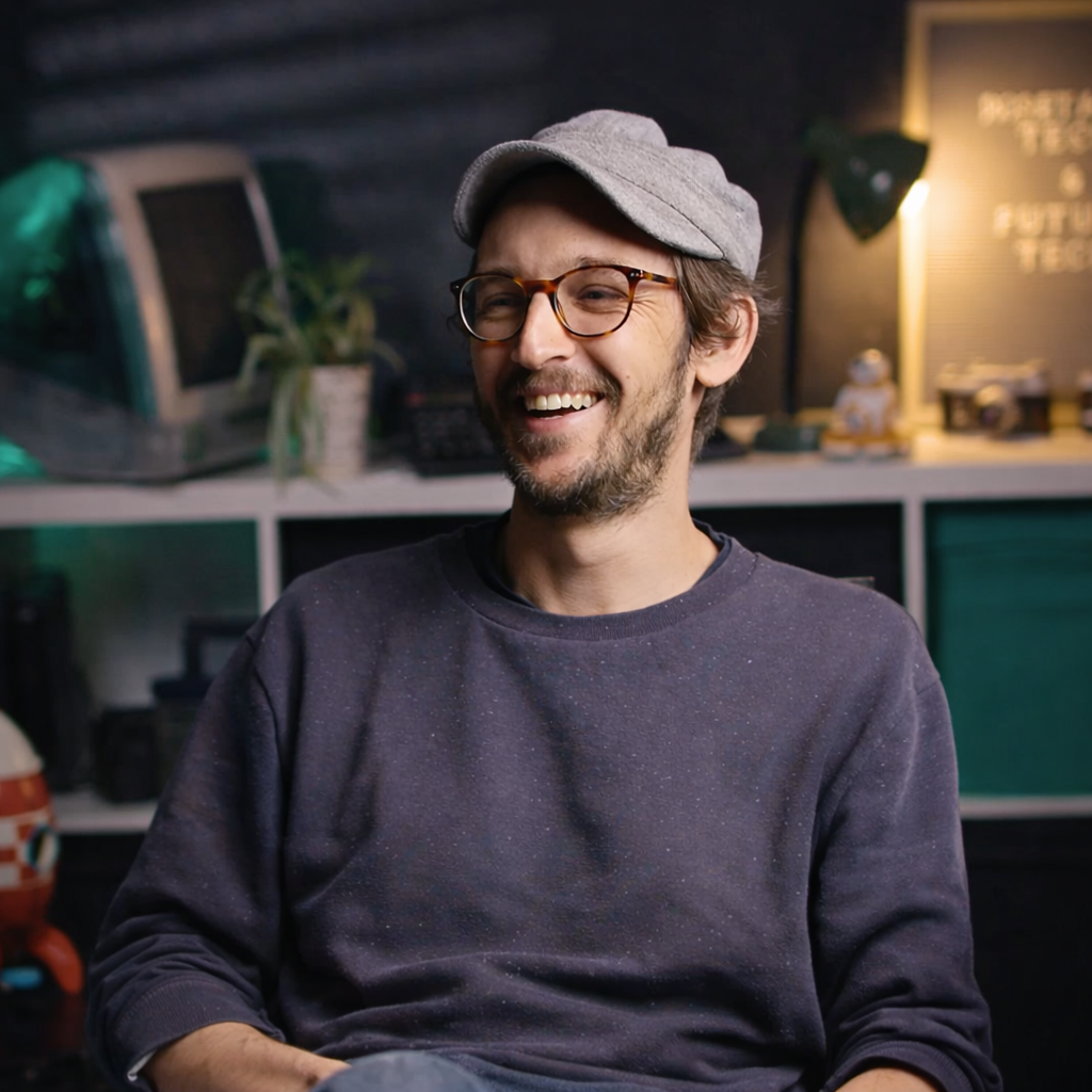 A man sitting in a modern, technology-themed room with a vintage computer on a shelf behind him. He is wearing a blue plaid shirt and sitting in front of a lightbox sign that reads, 'Nostalgic Tech & Future Tech.' The room has various tech gadgets and a plant in the background.
