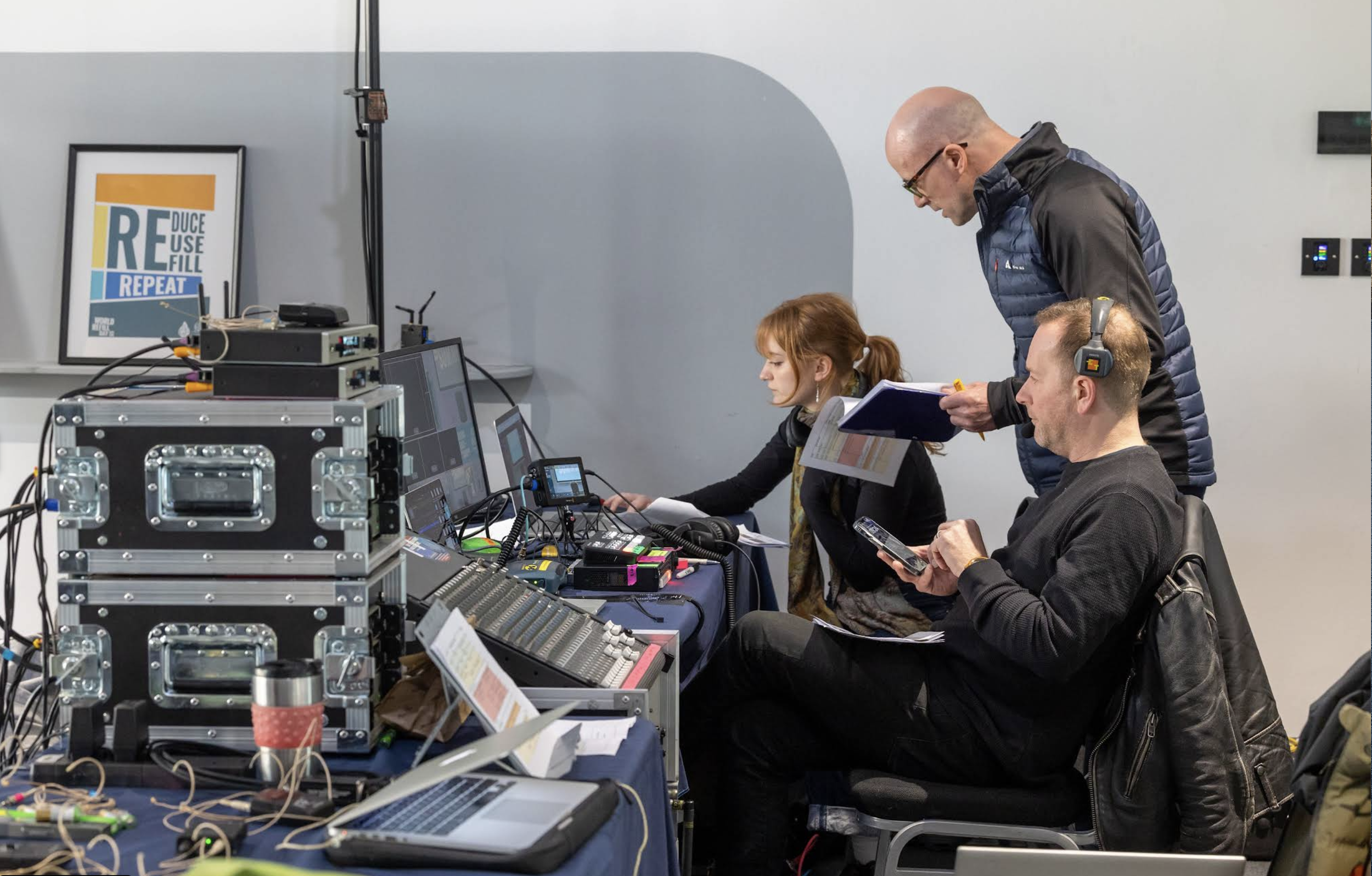 side-on view of 3 people sitting at a streaming setupp, looking at computers and clipboards
