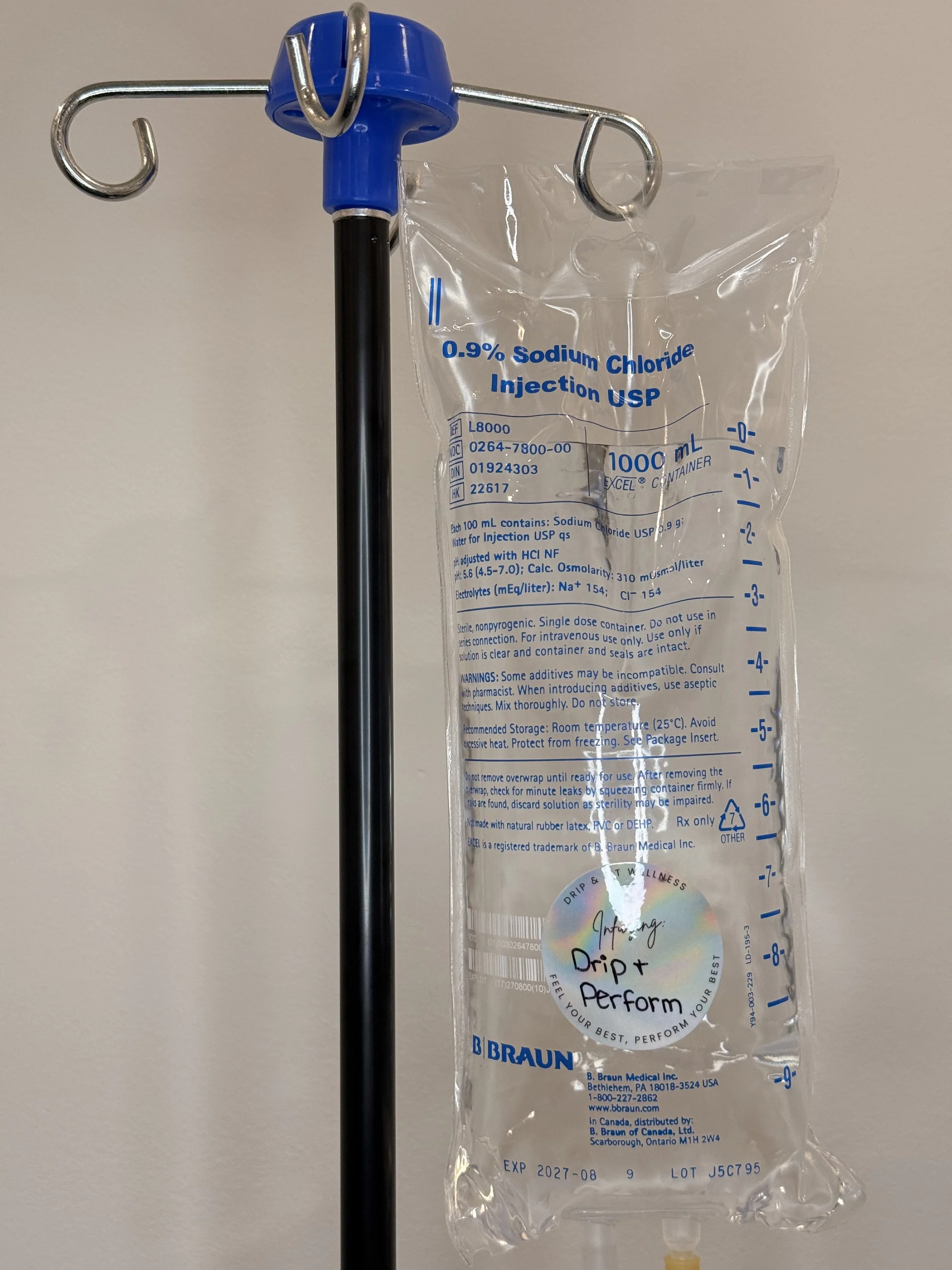 A medical IV bag labeled 0.9% Sodium Chloride Injection USP hanging on a black and blue IV pole. The bag has detailed text instructions, measurements, and a holographic sticker with the words "Infusing: Drip & Perform."