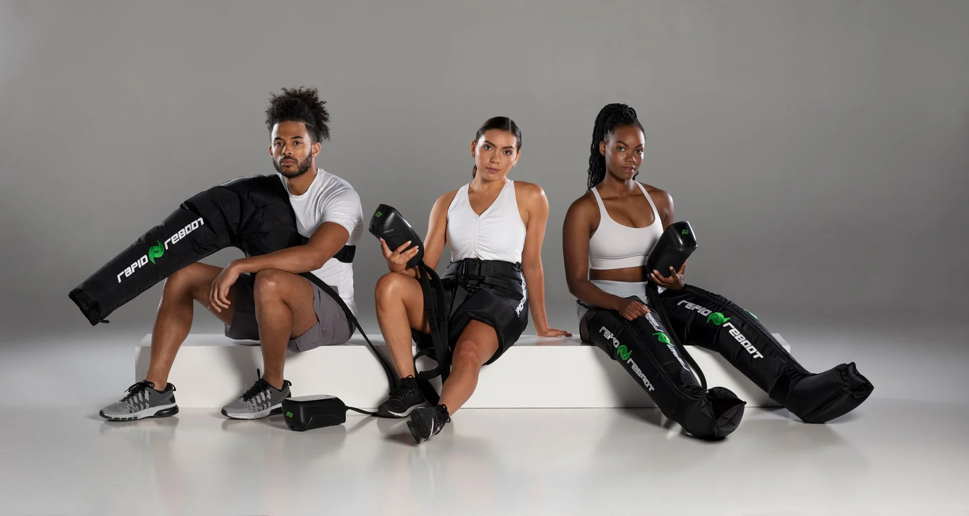 Three individuals seated on a white bench against a plain gray backdrop, each holding electronic fitness devices, with large black sports bags labeled 'Rapid Rebel' beside them.