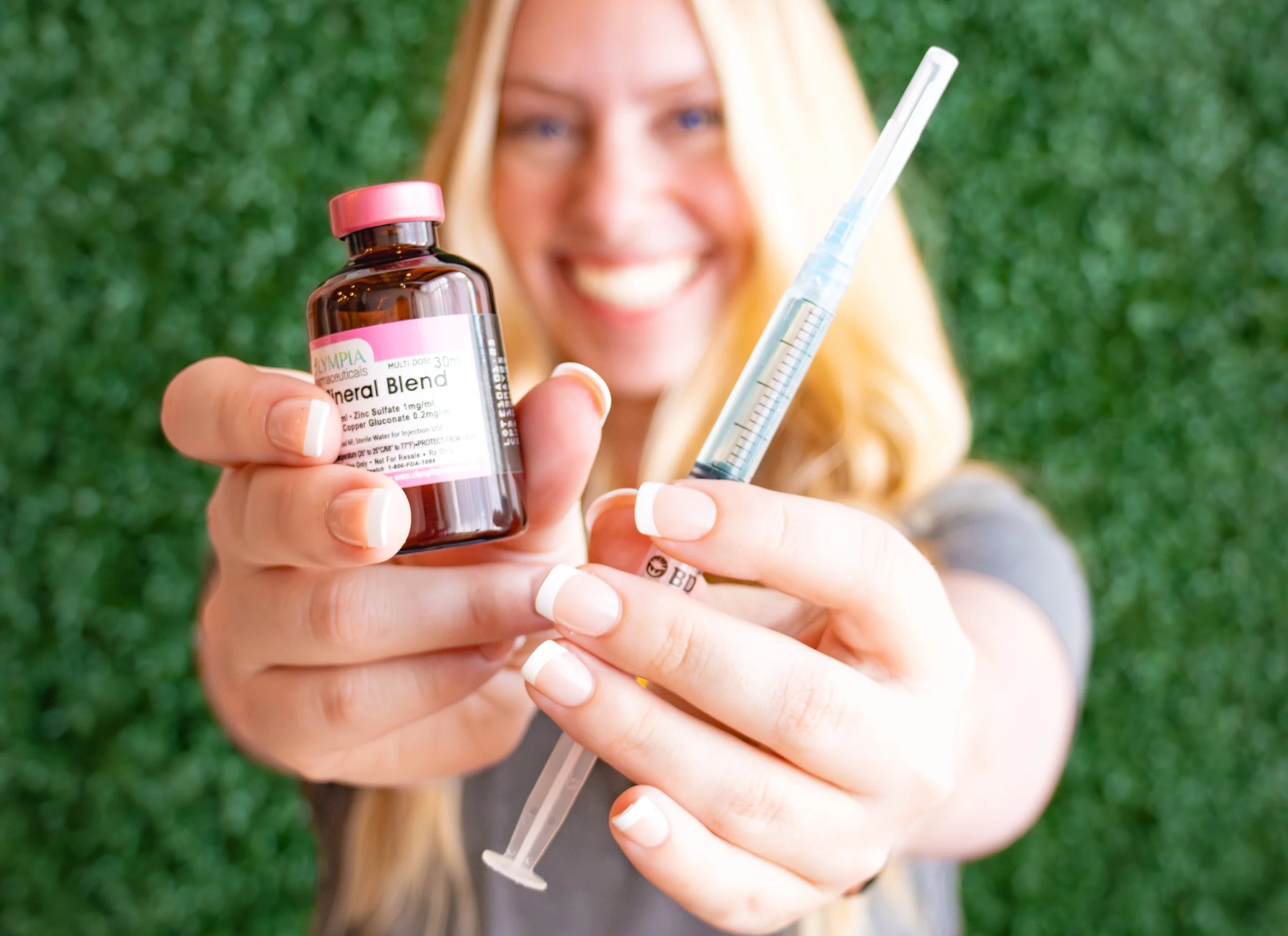 A woman smiling outdoors, holding a brown bottle labeled 'Mineral Blend' and a syringe, with a green background.