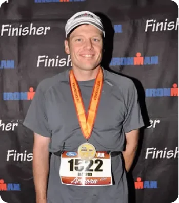 A smiling man wearing a gray athletic shirt, a striped cap, and a race bib with the number 1522 and the name 'Brad,' standing in front of a black backdrop with 'Finish' and 'Ironman' logos, and wearing a medal around his neck after a race.