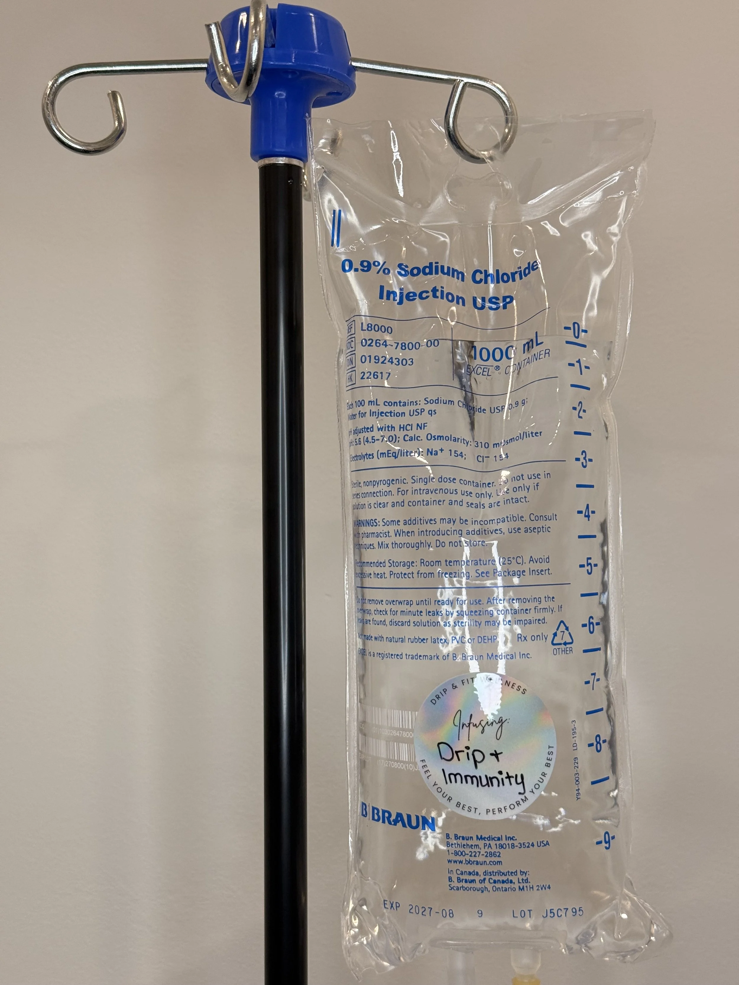 A clear plastic bag of 0.9% sodium chloride injection hanging from a metal IV pole with a blue top and hook, labeled for intravenous use, with measurement markings and a holographic sticker that reads 'Infusing: Drip & Immunity'.