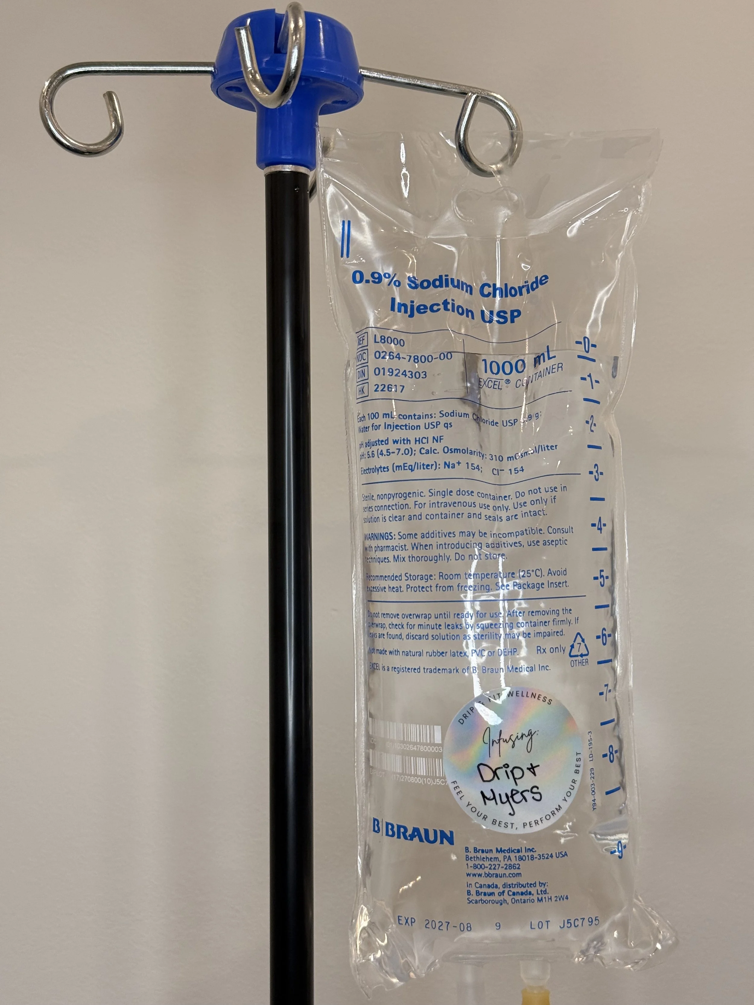 A medical IV fluid bag labeled '0.9% Sodium Chloride Injection USP' hangs on a metal IV pole. The bag is transparent with blue printed measurements, and the label indicates it contains 1000 mL of saline solution. There is a colorful holographic sticker on the bag with the text 'Infusing: Drip & Myers' and the phrase 'Feel your best, perform your best.'