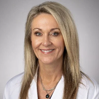 Headshot of a smiling woman with long blonde hair, wearing a white top and silver necklaces, against a neutral background.