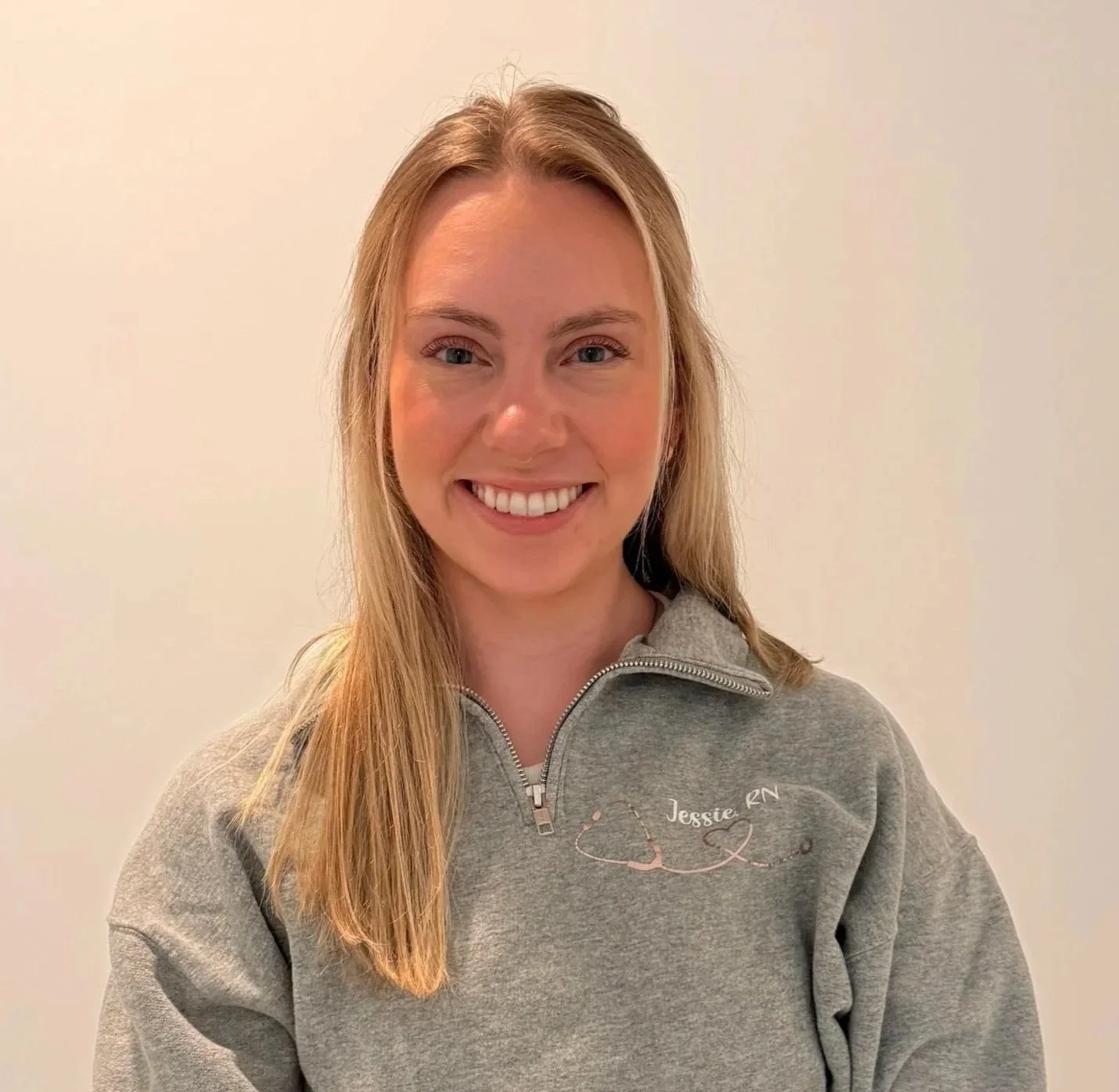 A smiling woman with long blonde hair, wearing a gray zip-up sweatshirt with a stethoscope and 'Jessie, RN' embroidered on it, standing against a plain white wall.