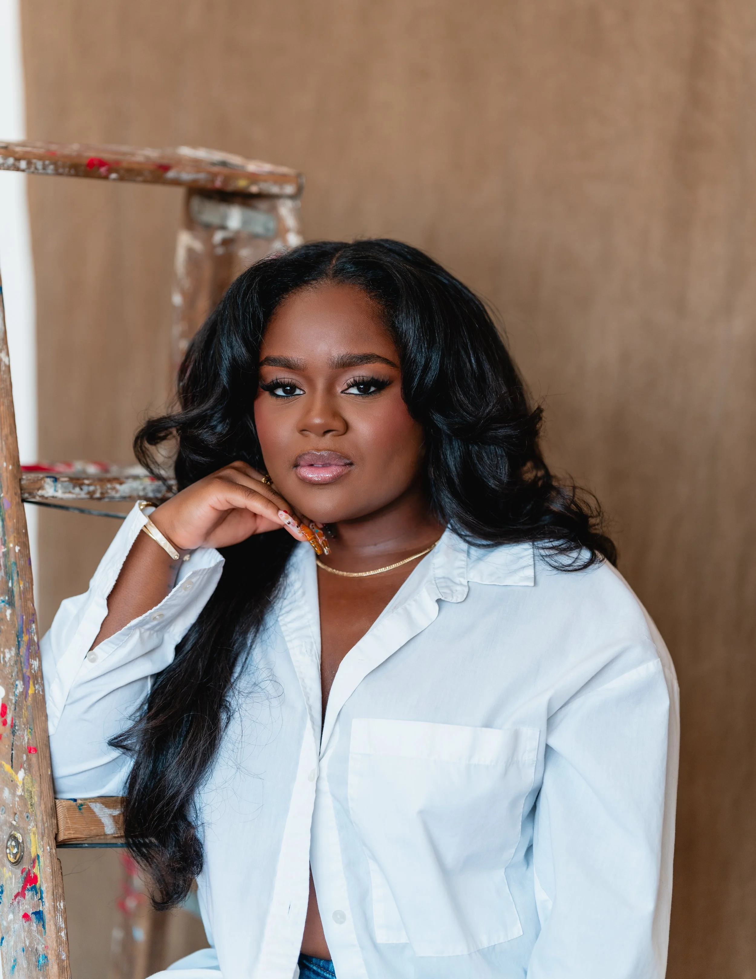 Portrait of a woman with dark hair and makeup, wearing a white shirt, standing next to a paint-splattered ladder in a studio setting.