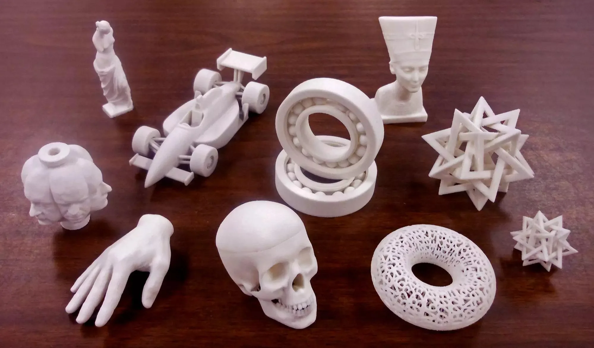 Collection of white 3D-printed objects including a hand, skull, geometric shapes, a race car, a statue, and an intricate donut shape