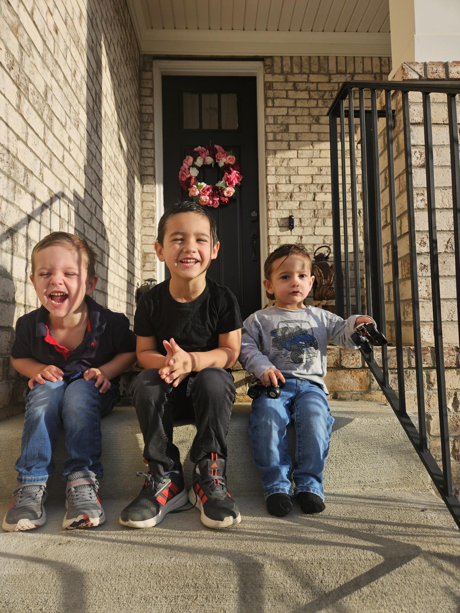 Three children sitting on a front porch with a brick wall and a floral wreath on the door in the background.