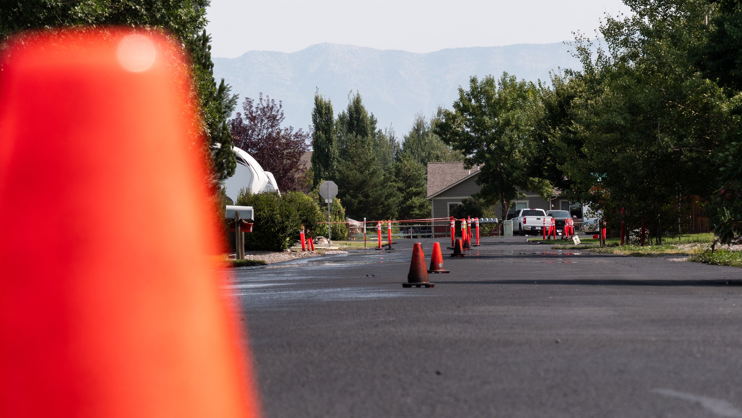 A street under construction with orange traffic cones and barriers, residential houses and trees in the background, mountains visible in the distance.