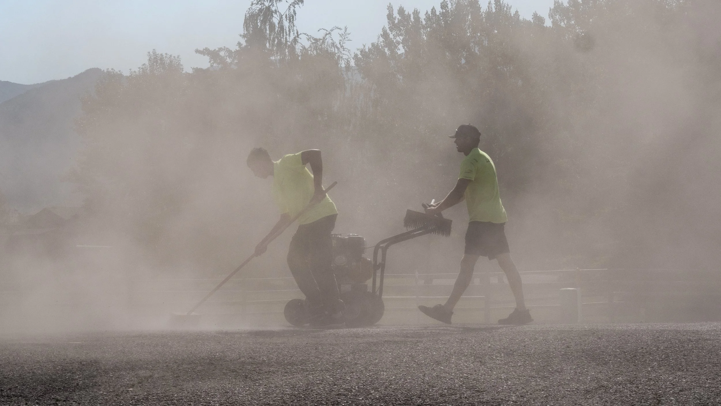 Two workers in bright yellow shirts using a machine to spray or clean a dusty or dirty surface, with dust or mist in the air and trees in the background.
