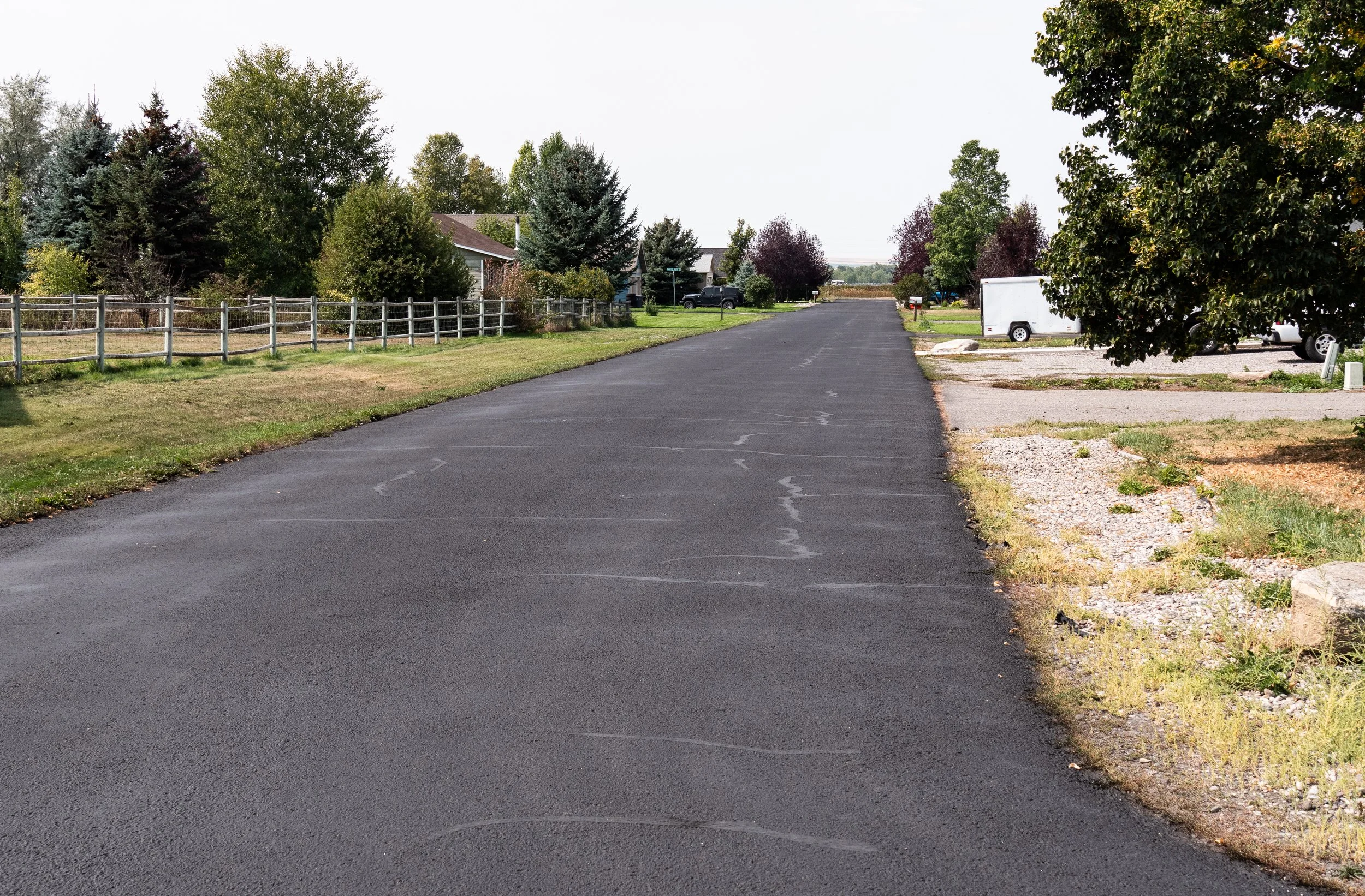 A suburban street with freshly paved asphalt, grass lawns, trees, and parked vehicles along the side.