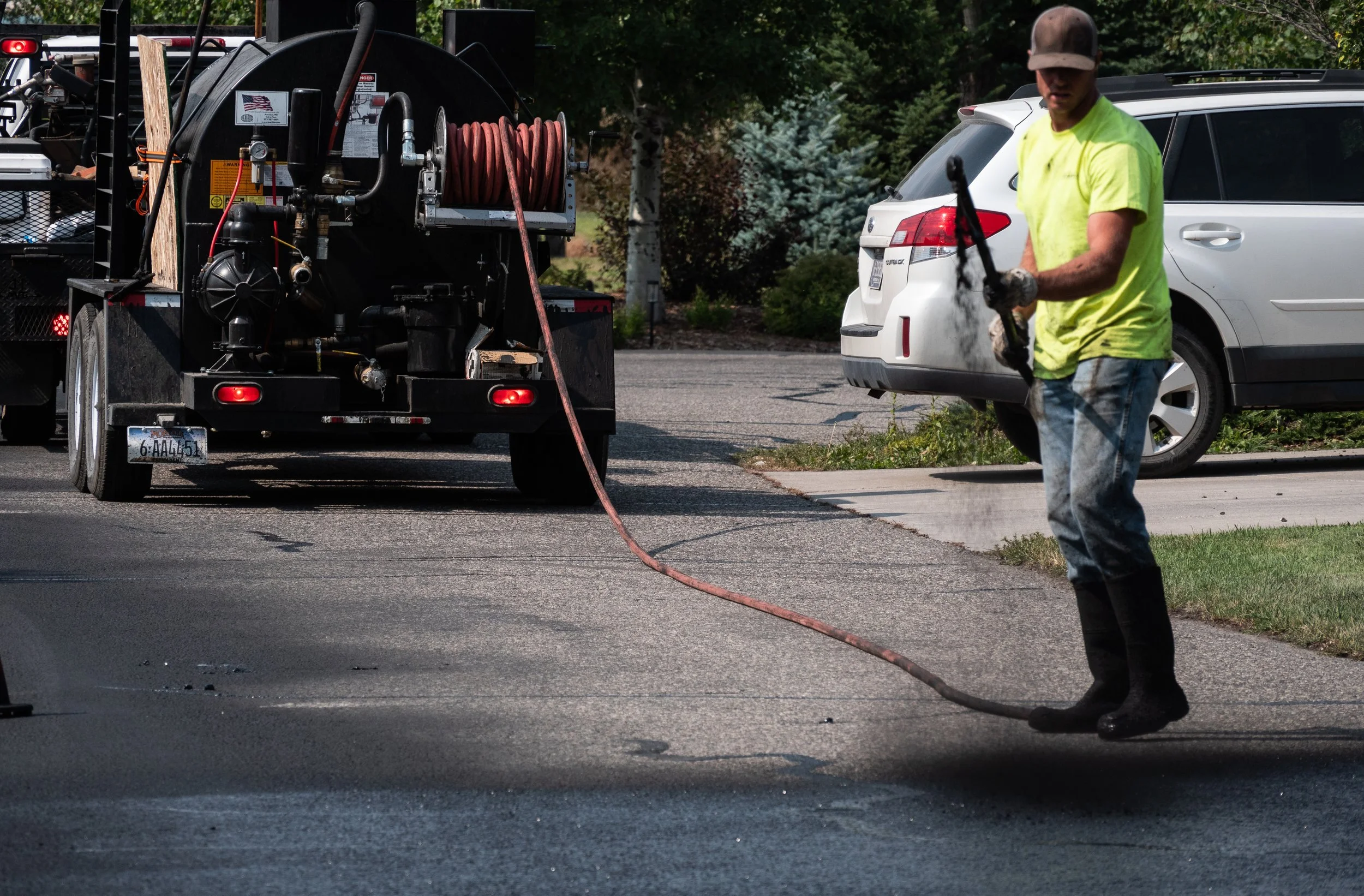 Worker in yellow shirt and boots using a high-pressure hose to clean a street in a residential neighborhood.