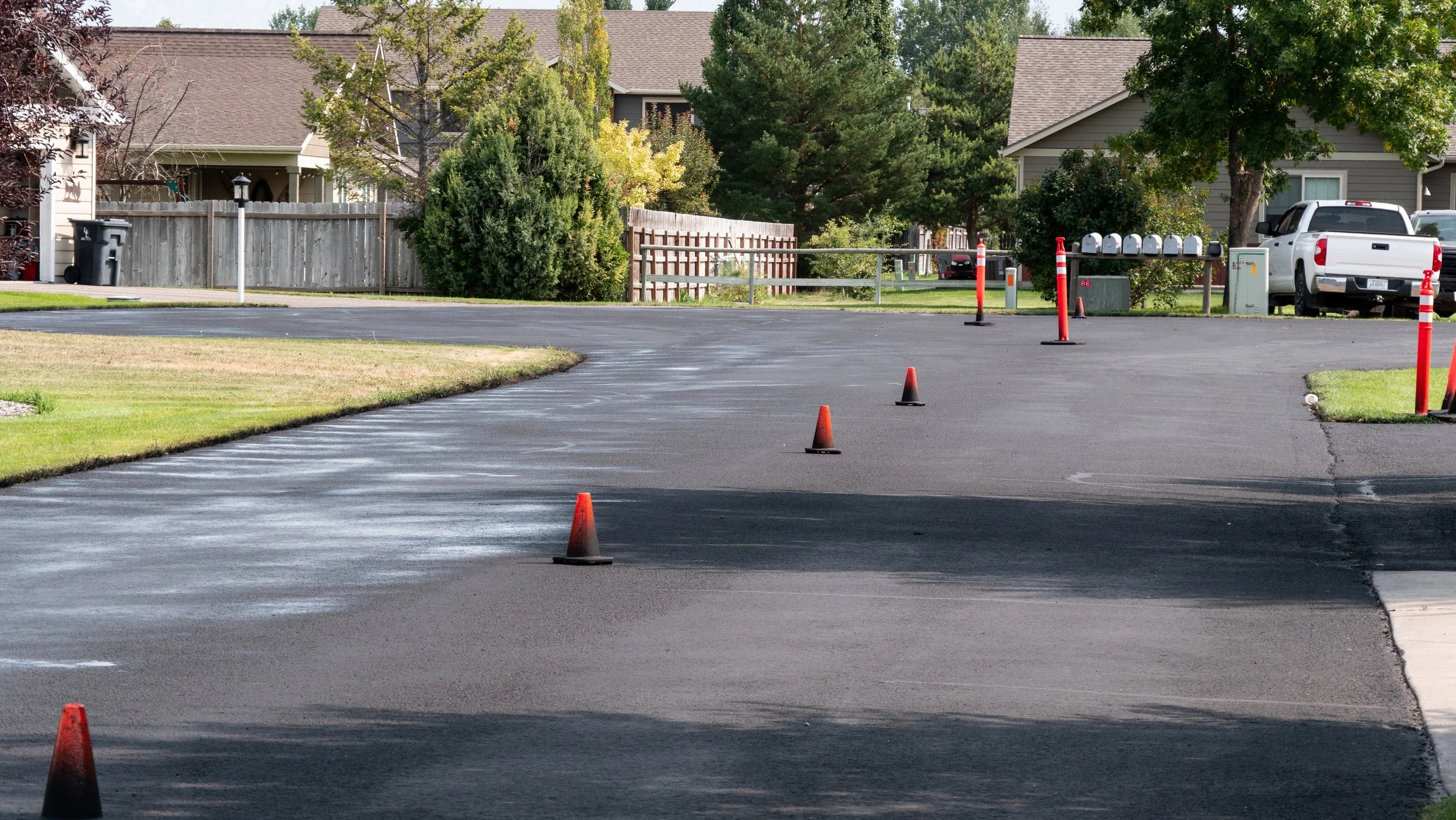 A freshly paved asphalt driveway with orange traffic cones lining a section of it, adjacent to a grassy lawn and residential houses in the background.