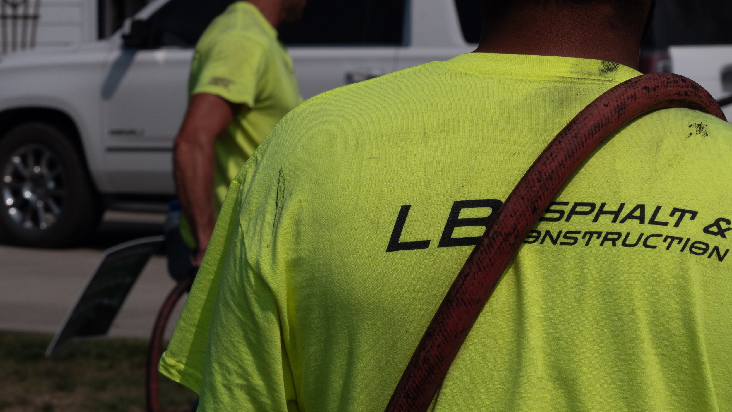 Close-up of a construction worker in a bright yellow shirt with the text 'LB ASPHALT & CONSTRUCTION' on the back, standing outdoors near construction equipment and a white vehicle.