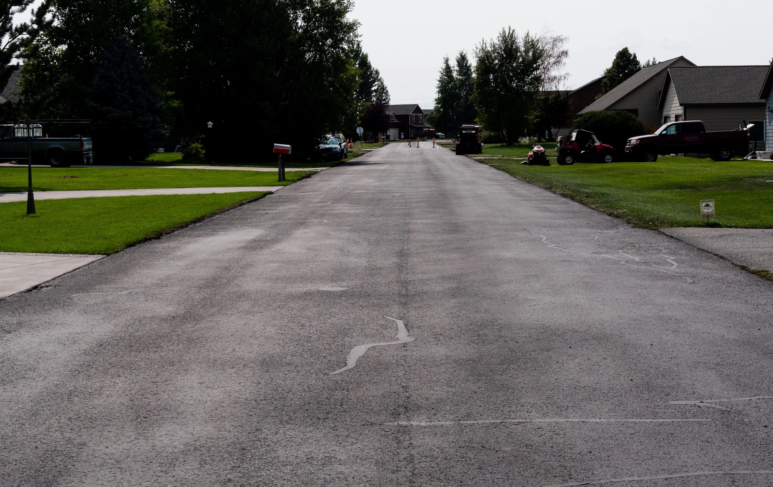 A residential neighborhood street with grass lawns, trees, houses, and parked trucks and cars, some orange traffic cones at the far end of the street.