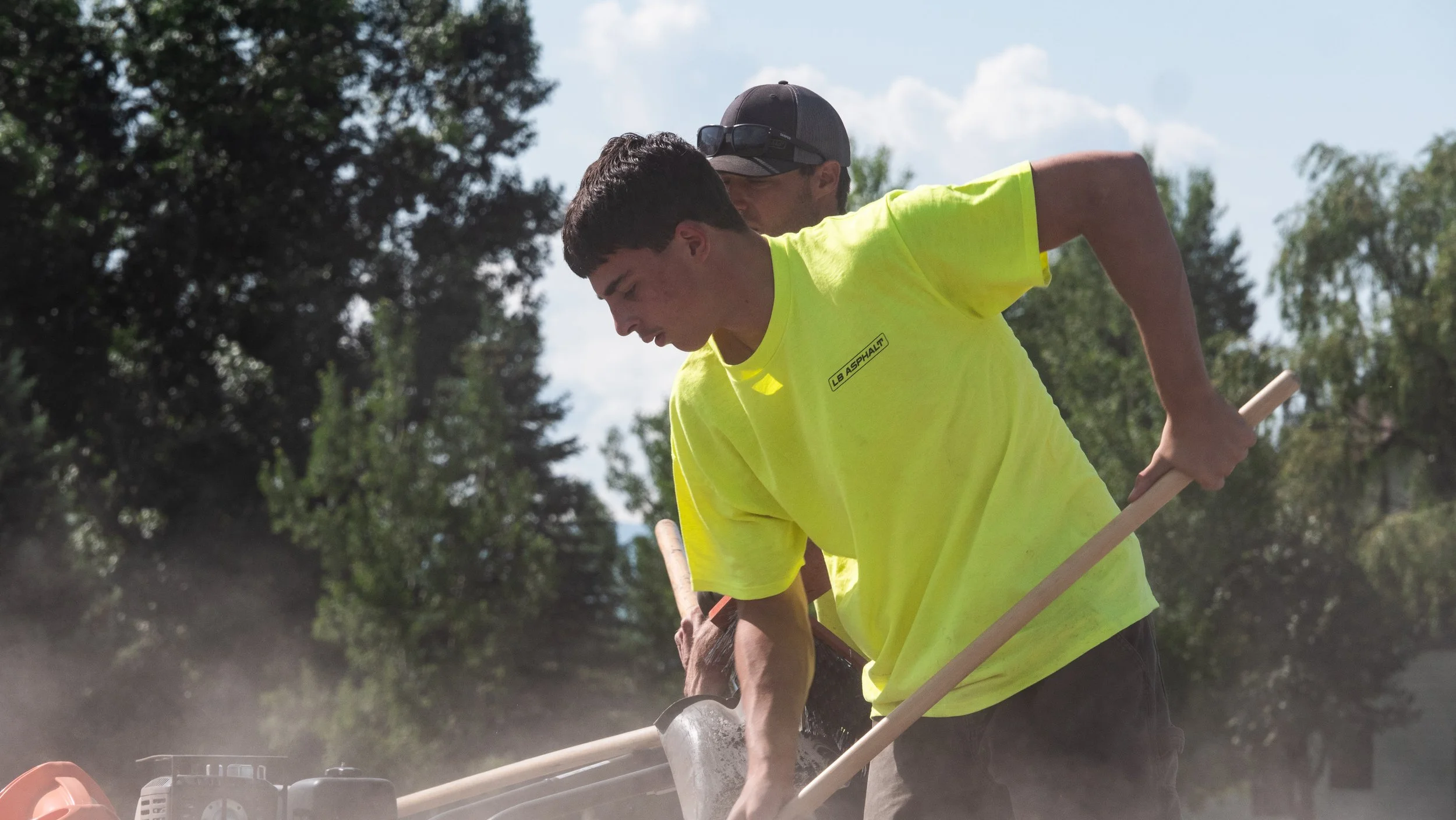 Two young men working outdoors, one wearing a bright yellow shirt and the other a grey cap, using tools near a construction or landscaping site with trees in the background.