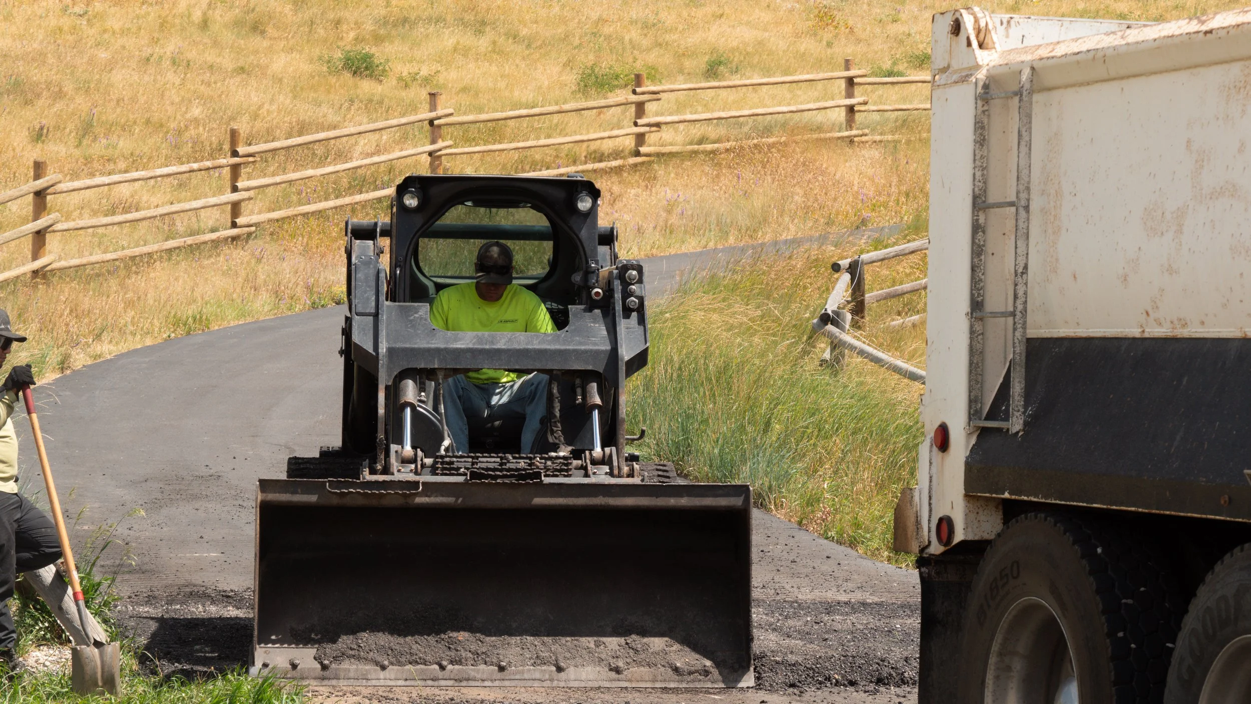 A construction worker operating a skid-steer loader on a rural road, with another worker shoveling gravel nearby and a pickup truck on the right, with a grassy hill and wooden fence in the background.