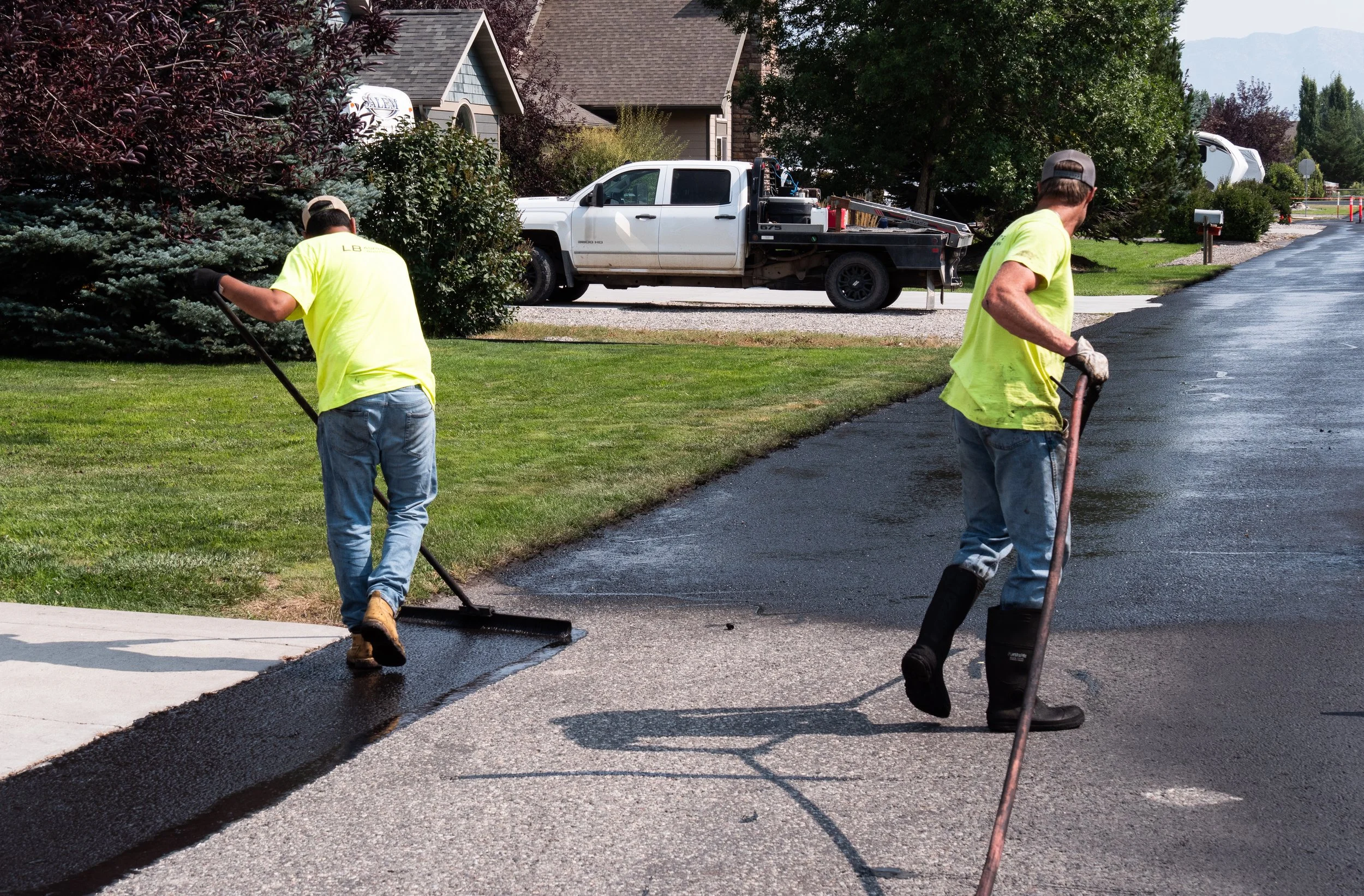 Two workers in neon yellow shirts and jeans are paving a residential street with asphalt. One worker is spreadng asphalt near the sidewalk, while the other is smoothing the asphalt with a longer tool. A white truck with tools and equipment is parked 