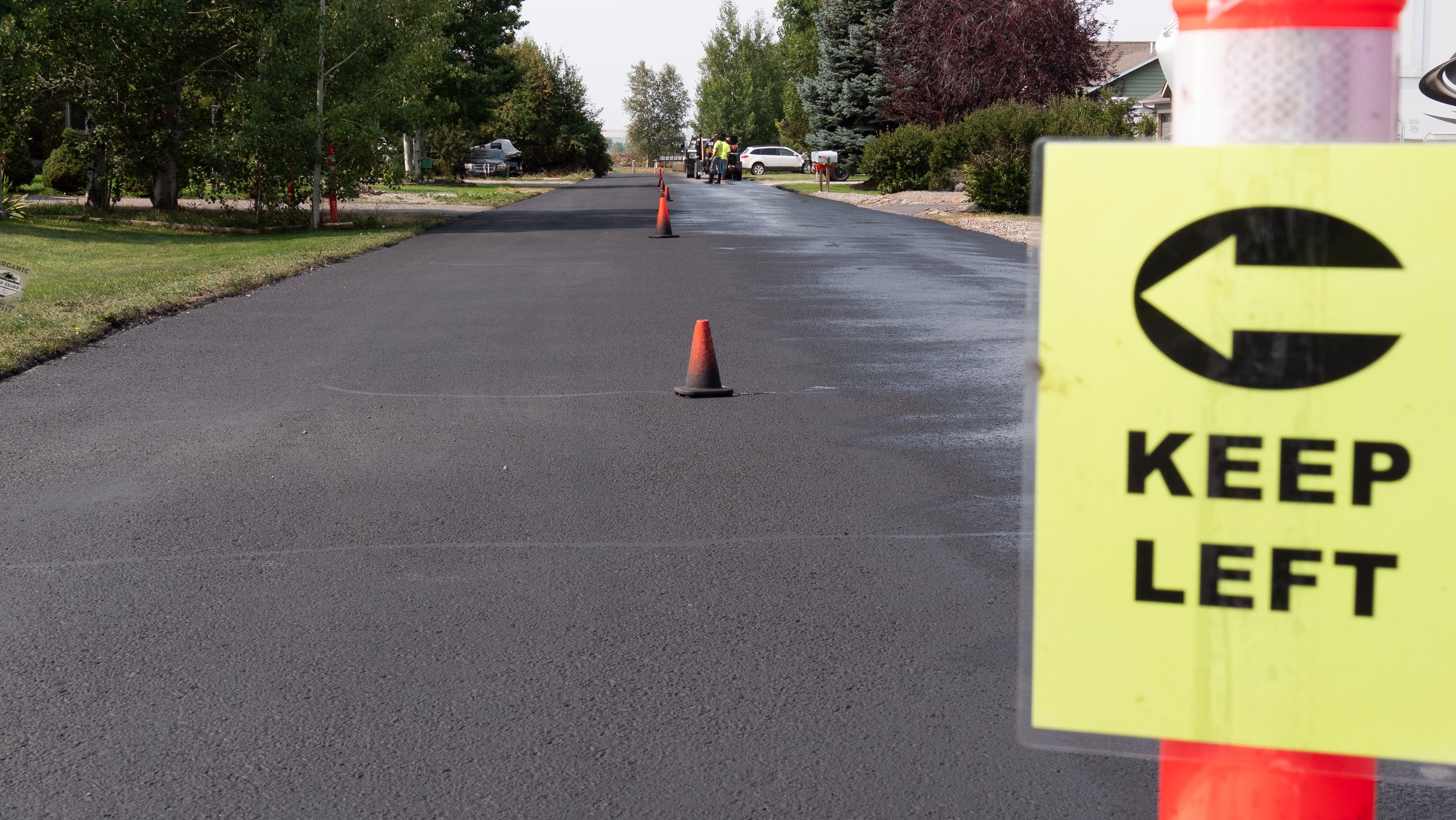 Freshly paved asphalt street with orange traffic cones and a yellow sign that reads "Keep Left" with a left arrow, in a suburban neighborhood with trees and houses.
