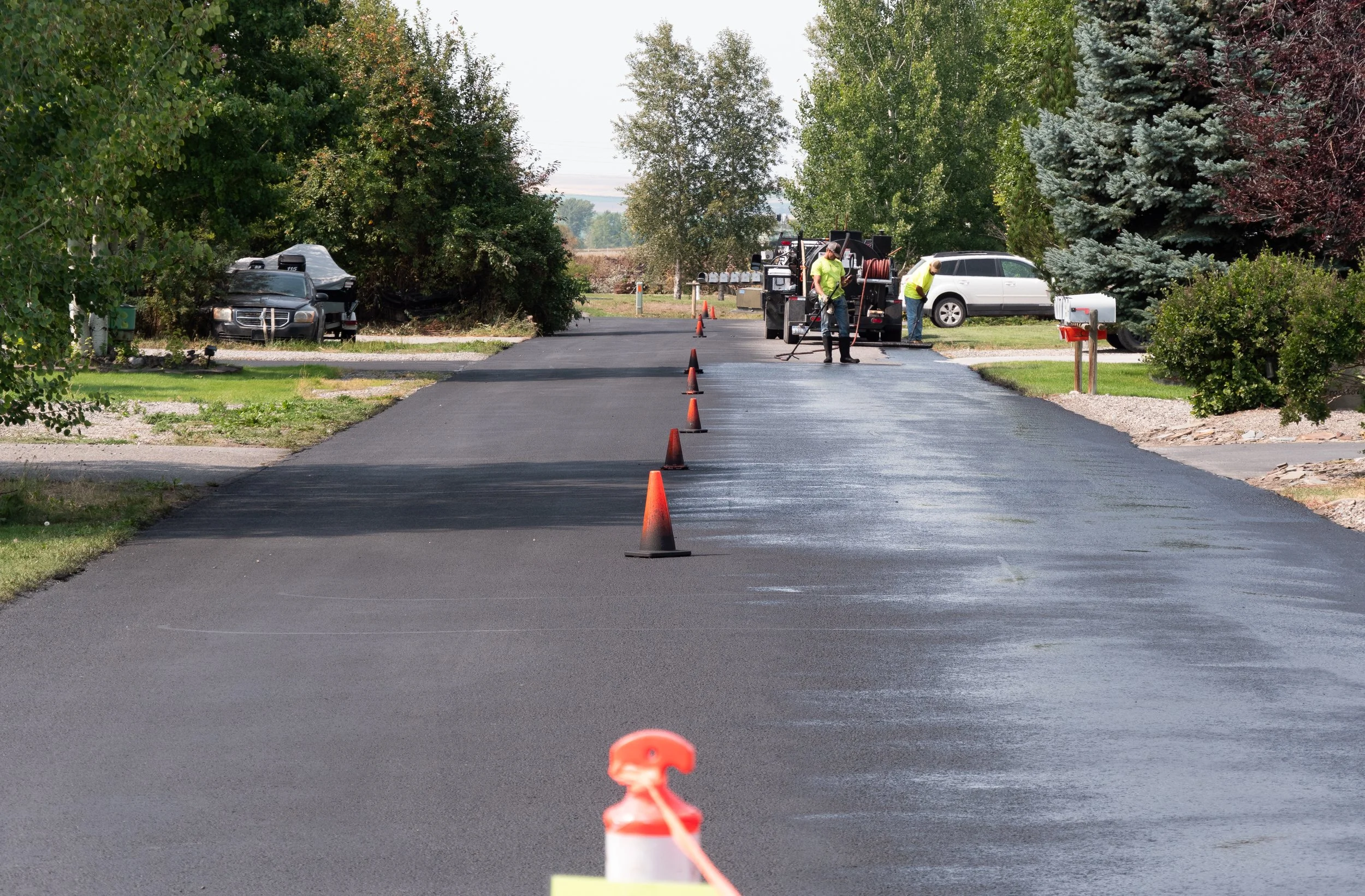 Workers paving a residential street with fresh asphalt, orange traffic cones marking the work area, and trees lining the street, with parked cars on either side.