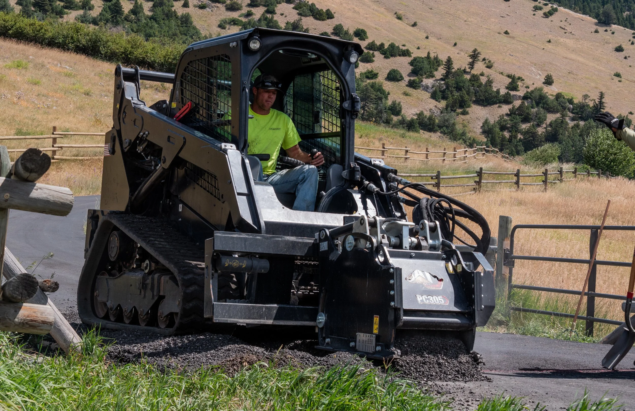 A man in a neon green shirt operates a black compact track loader with a front blade, fixing asphalt on a rural road surrounded by grassy fields, fences, and hills in the background.