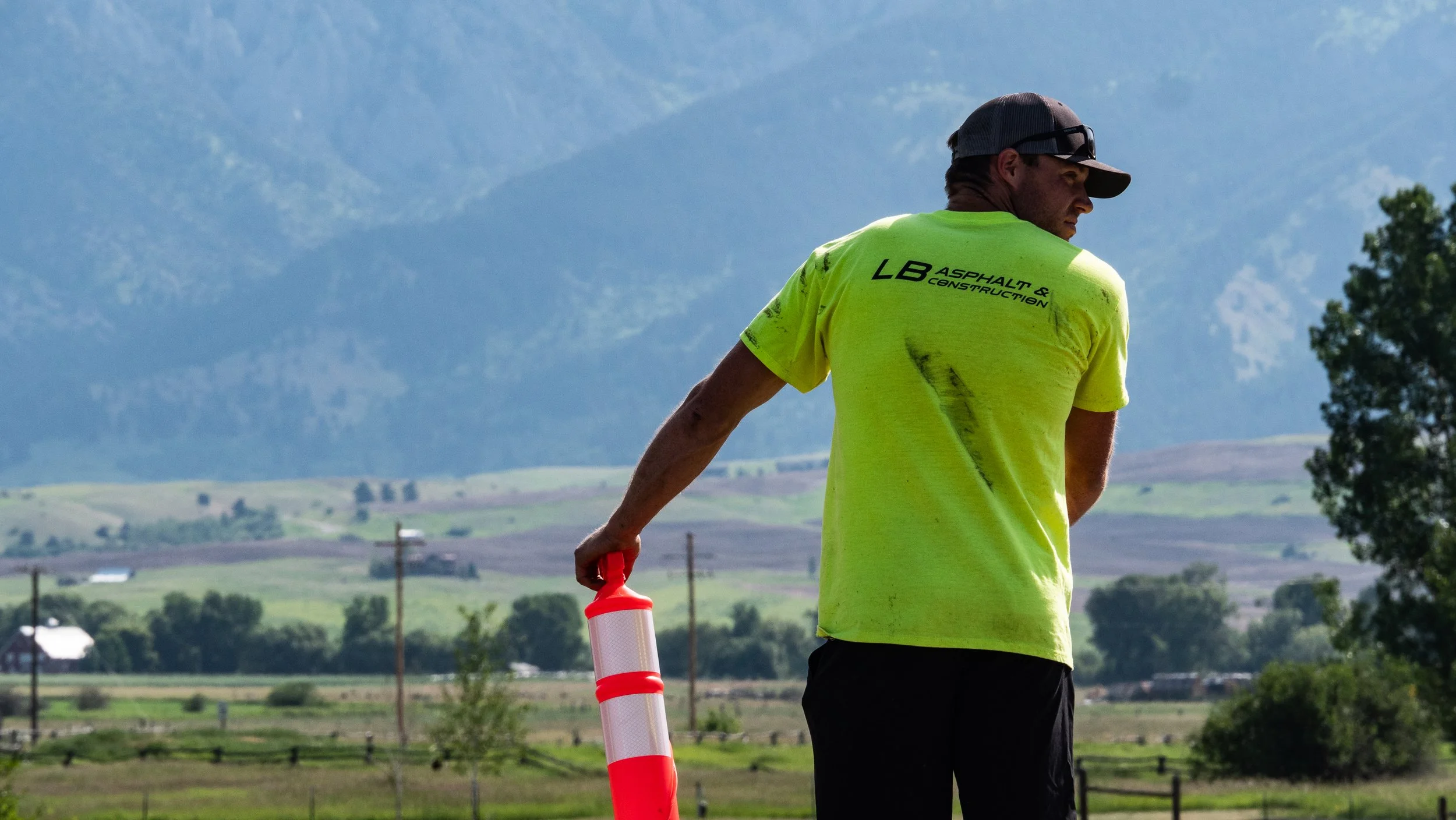 A man in a yellow T-shirt and cap holding a striped traffic cone, standing outdoors with mountains and farmland in the background.