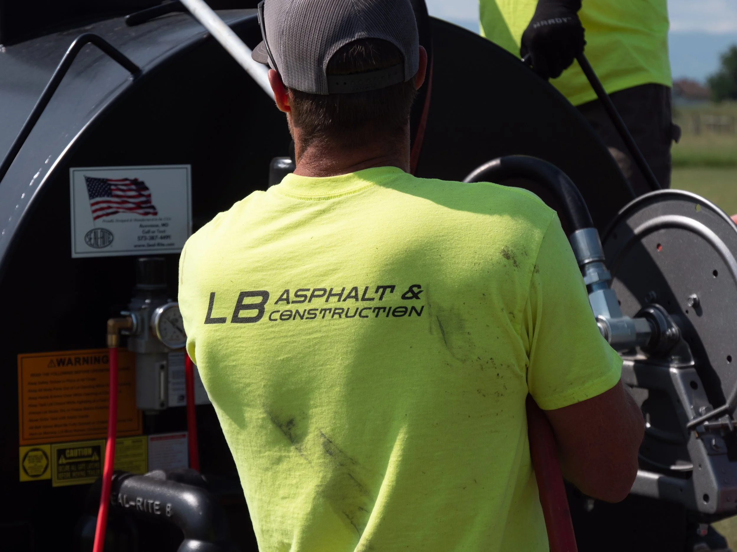 Worker in a neon yellow shirt with 'LB Asphalt & Construction' written on the back, working on a piece of construction equipment outdoors during the day.