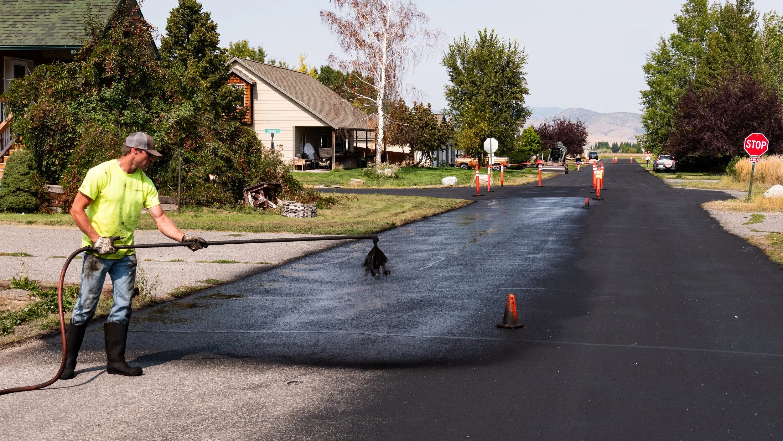 A construction worker spraying asphalt on a street using a spray gun, with traffic cones and barriers to block off the area.