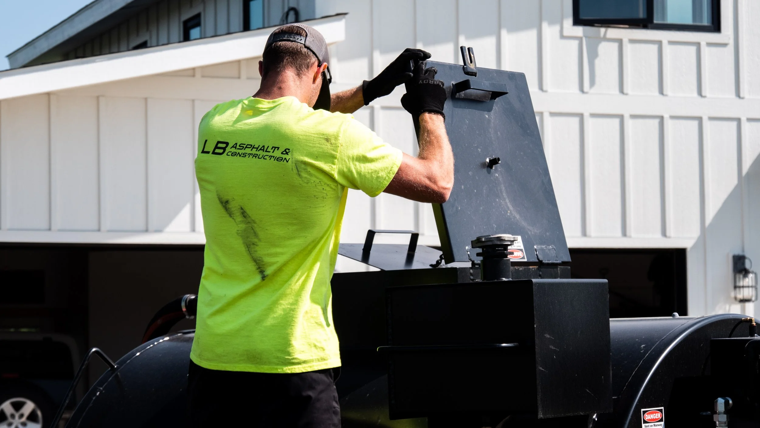 A worker in a bright yellow shirt with the logo 'LB Asphalt & Construction' is operating a black industrial machine outside a residential building with white siding. The worker is wearing black gloves and a gray cap, facing away from the camera.