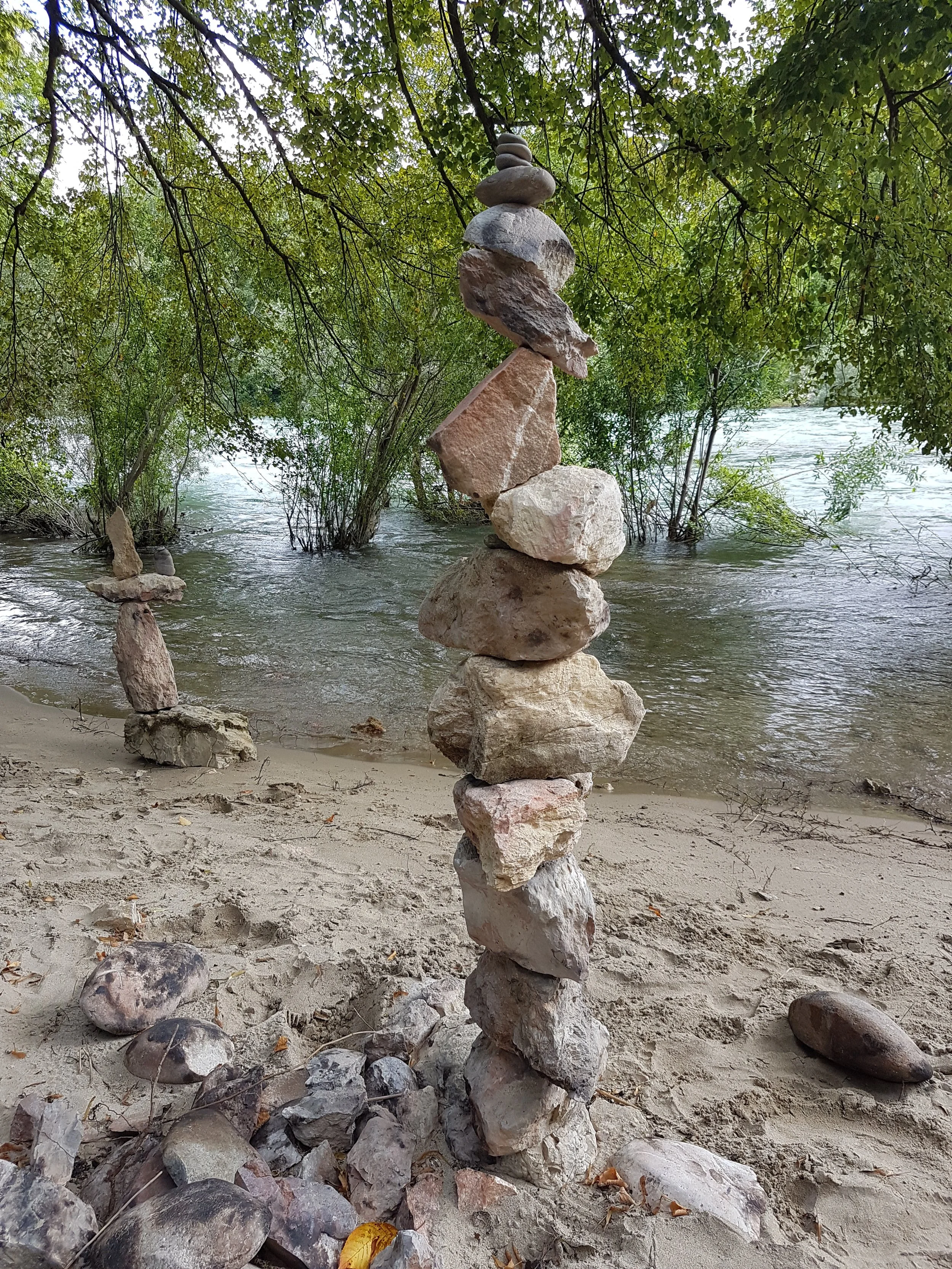 Ein Strand mit einem Steinturm, der aus mehreren Steinen besteht, vor einem Wasserlauf und umgeben von Bäumen.