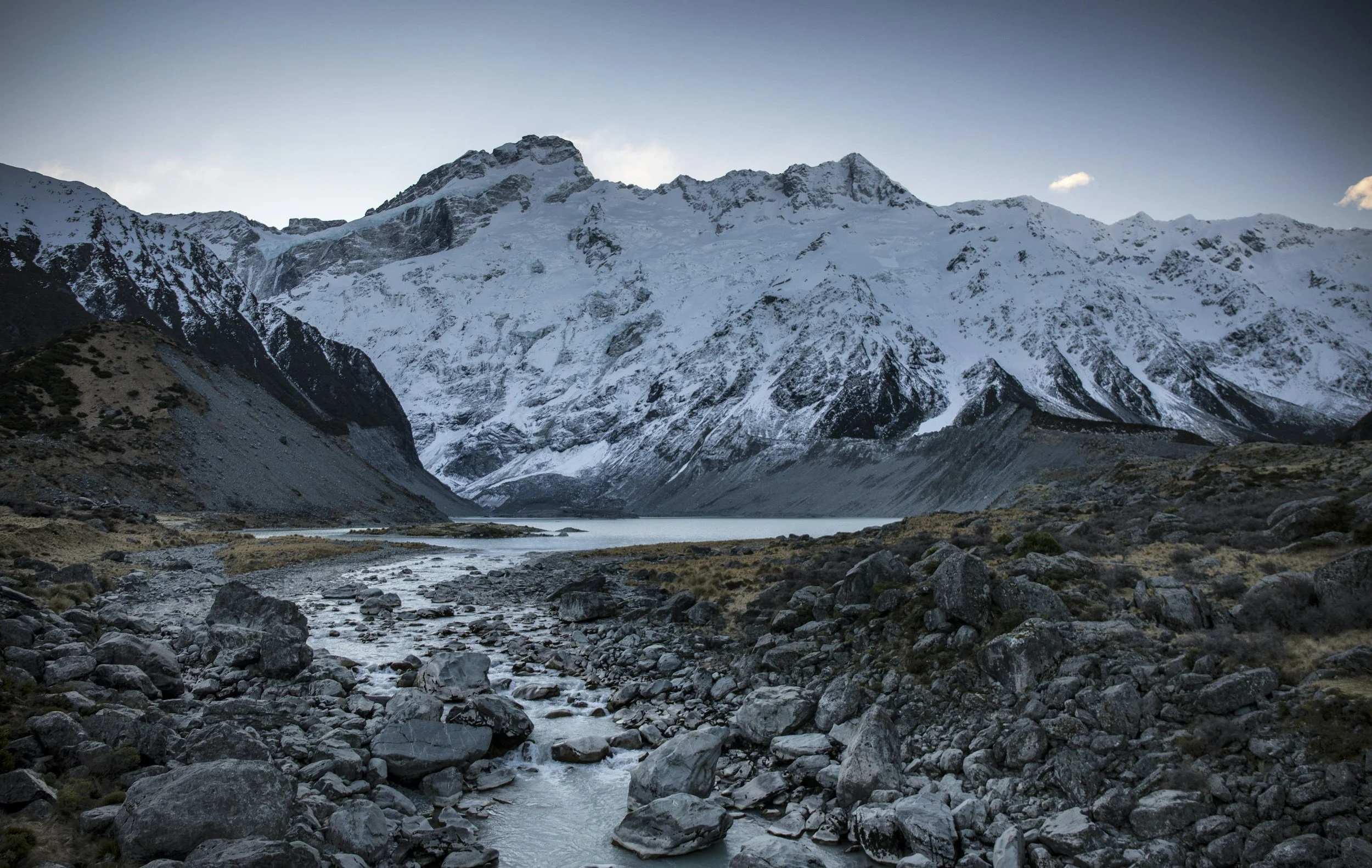Berglandschaft mit schneebedeckten Gipfeln, einem See im Tal und einem Fluss, der über Gestein fließt.