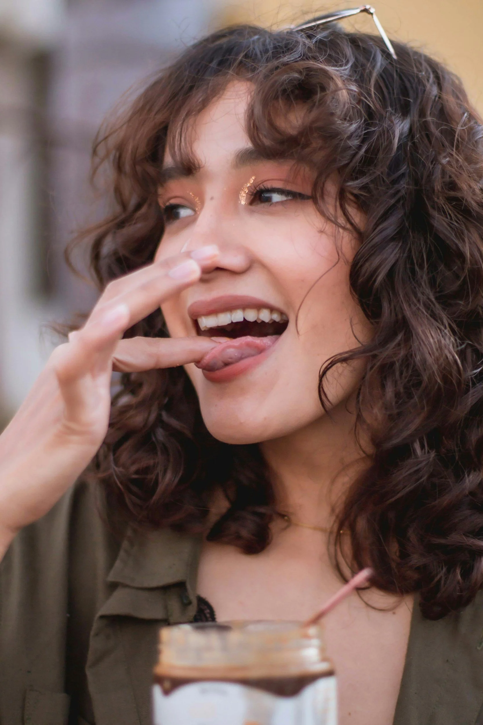 Junge Frau mit lockigen Haaren und Glitzer im Gesicht, leckt am Finger, auf einem Tisch steht ein Glas mit einem Strohhalm.