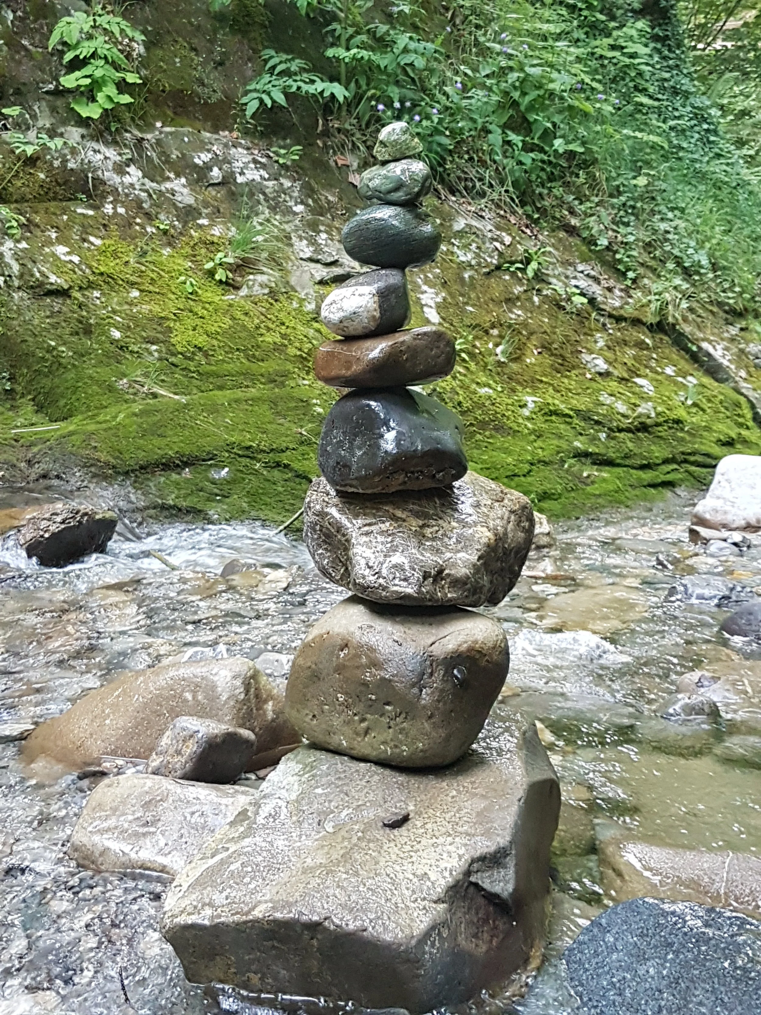 Ein Steinturm aus mehreren übereinander gestapelten Steinen in einem Bach mit grüner Ufervegetation im Hintergrund.