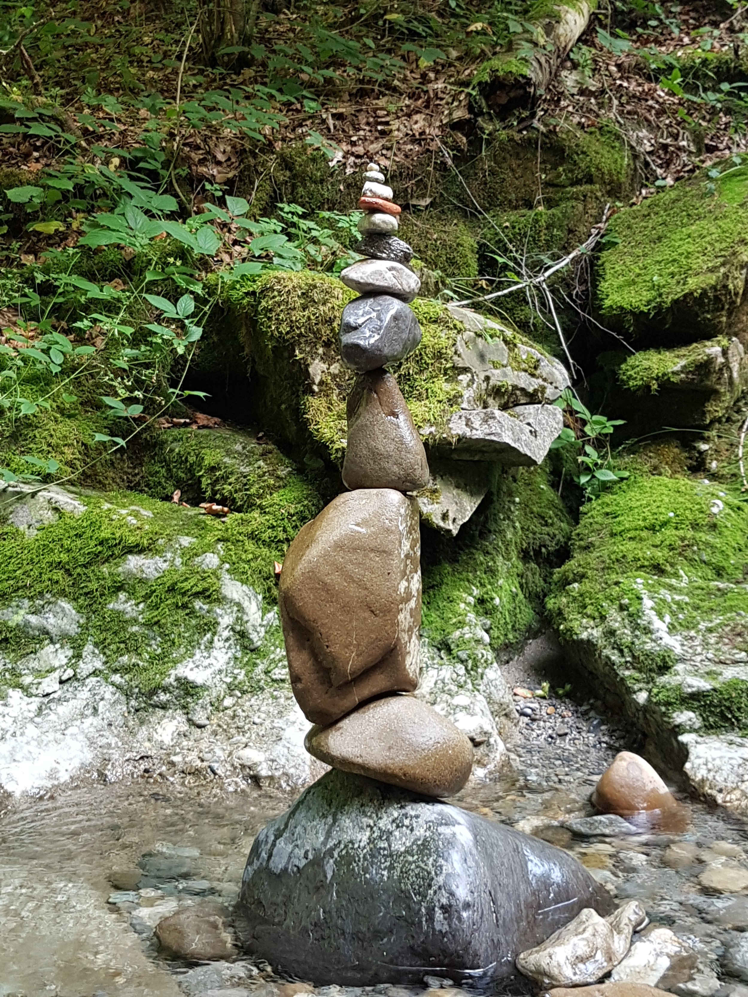 Steinmandala in einem Moos- und Laubwald an einem Bach, mit mehreren gestapelten Steinen unterschiedlicher Formen und Größen.