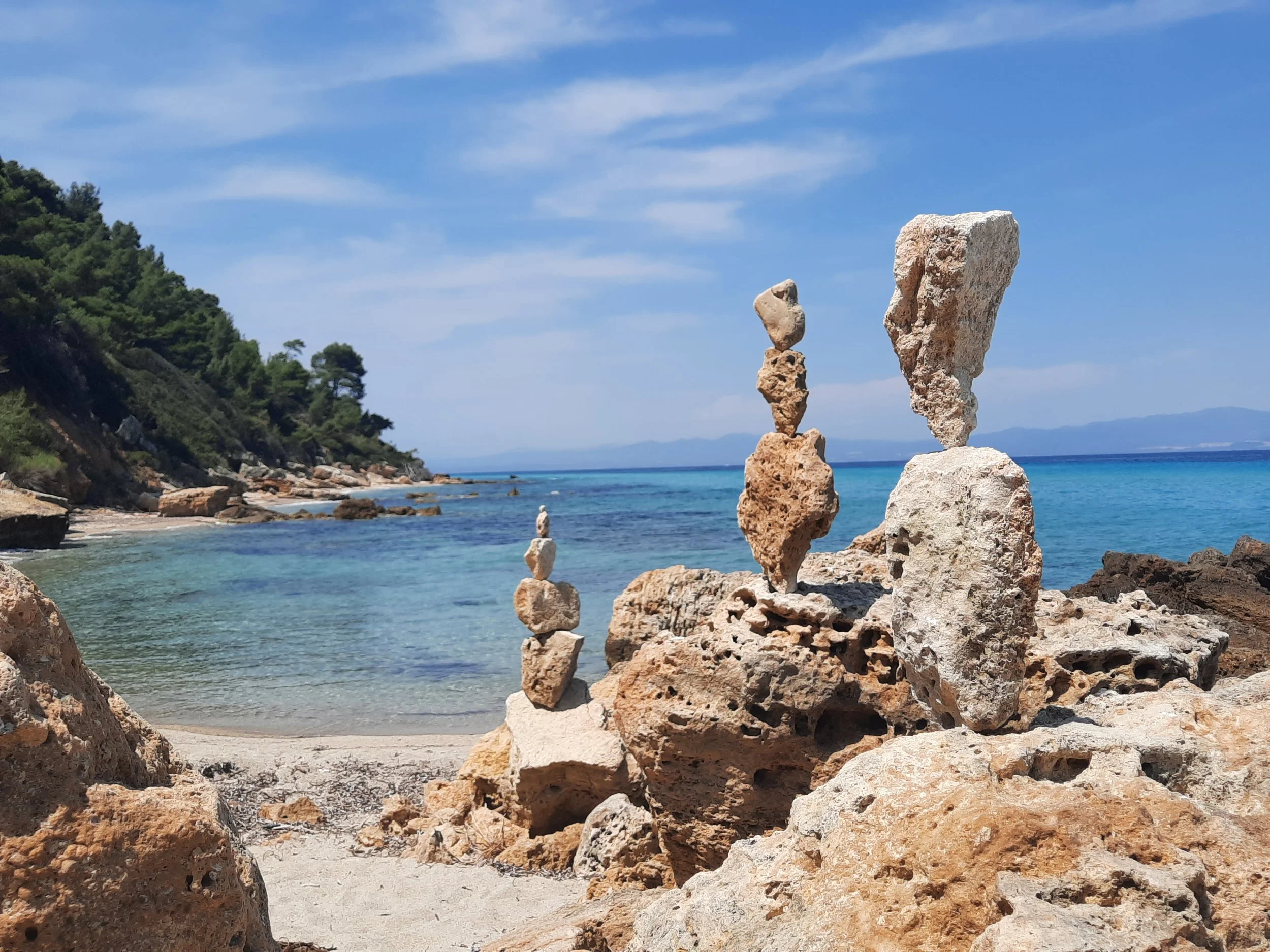Blick auf einen Strand mit Felsen und in den Himmel aufgestapelte Steine, im Hintergrund das Meer und eine bewaldete Küstenlinie bei sonnigem Wetter.