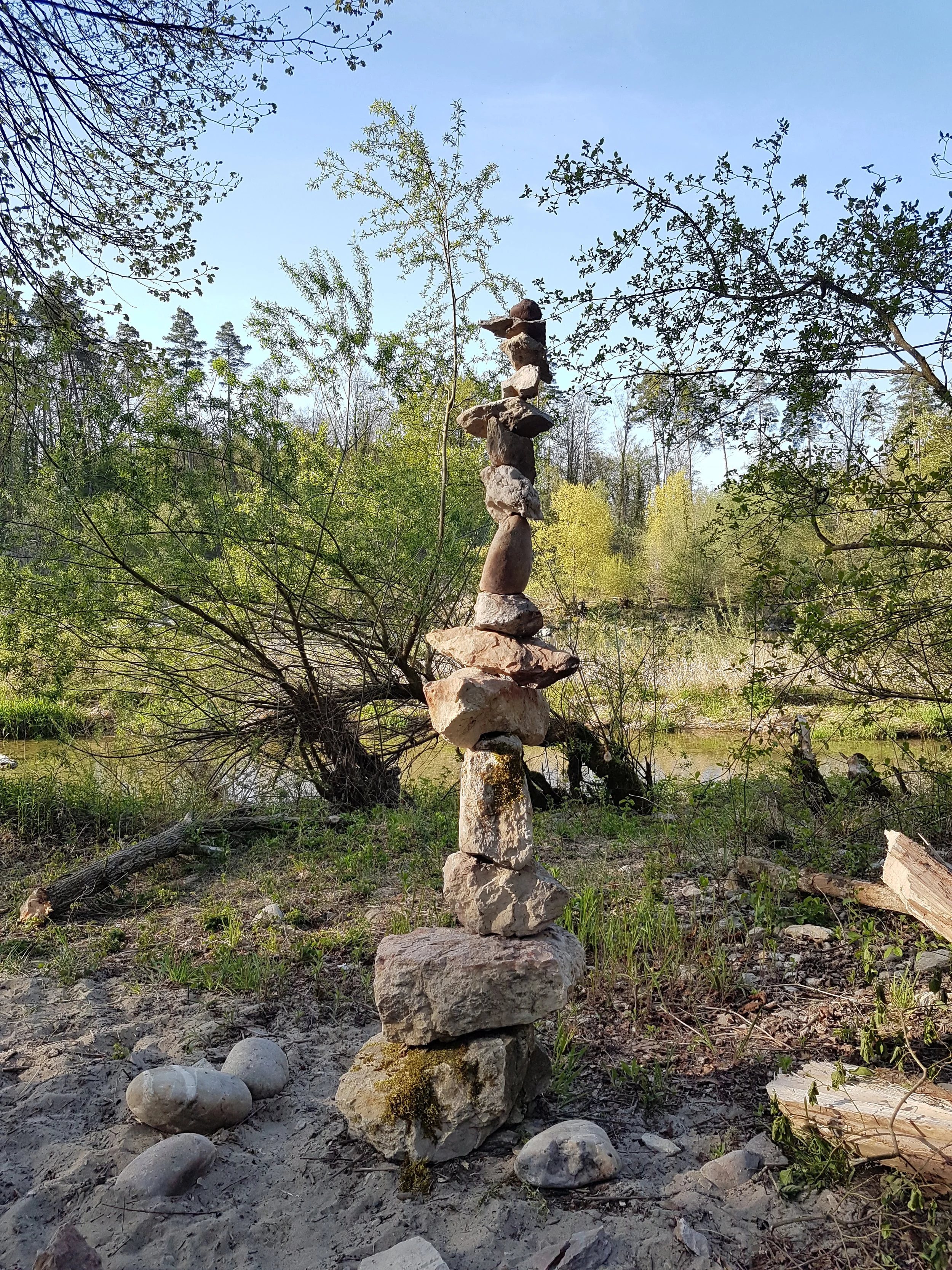 Steinmännchen aus verschieden großen Steinen in einer natürlichen Umgebung mit Bäumen und Wasser im Hintergrund, bei Sonnenlicht aufgenommen.