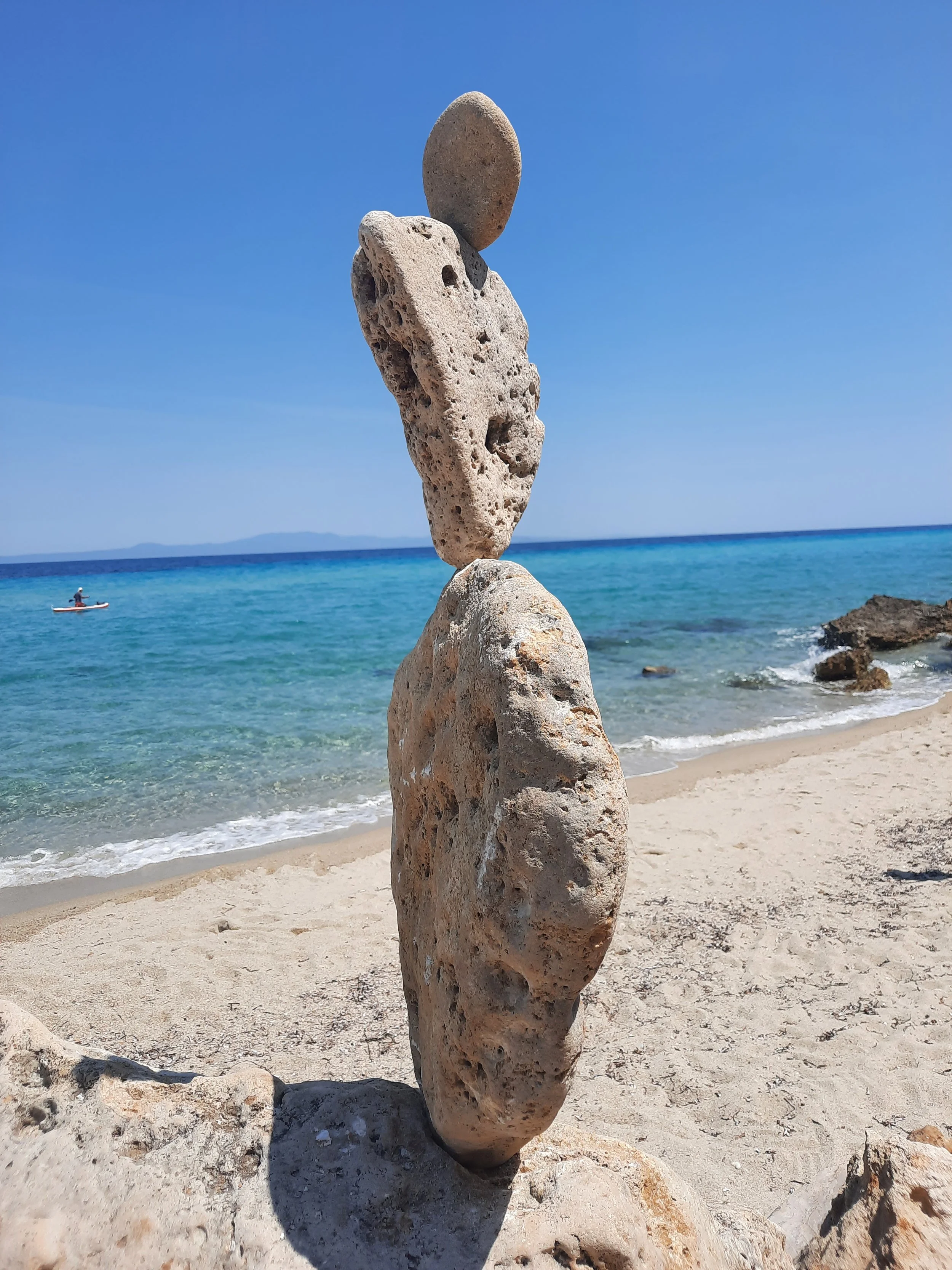 Fünf Steine, die auf einem Strand übereinander gestapelt sind, mit Blick auf das Meer und einen blauen Himmel.