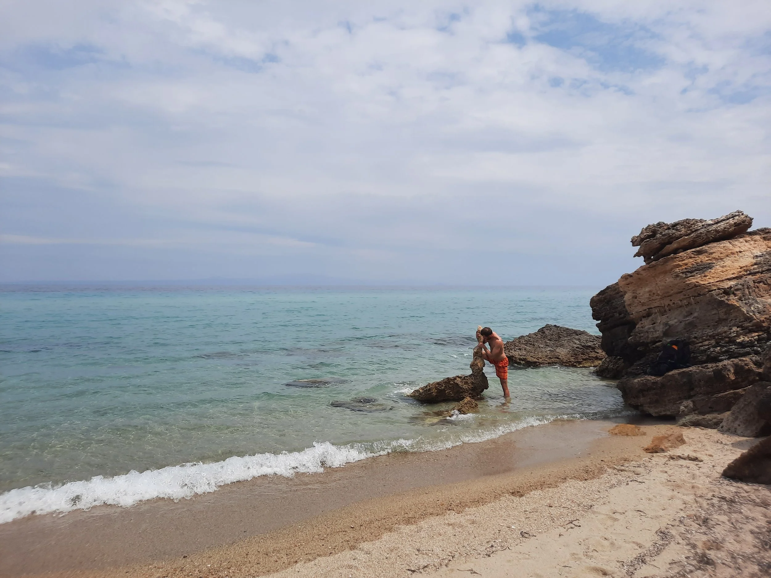 Ein Mann in roten Badeklamotten steht in flachem Wasser am Strand, umgeben von Felsen, mit einer leichten Wolkenbedeckung am Himmel.