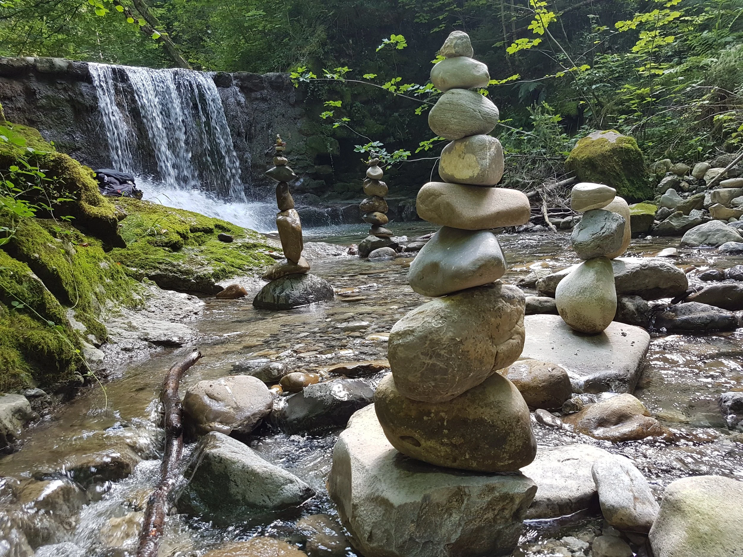 Ein Bach mit mehreren aufeinandergestapelten Steinen im Vordergrund, ein kleiner Wasserfall im Hintergrund, umgeben von grünen Bäumen und Moos