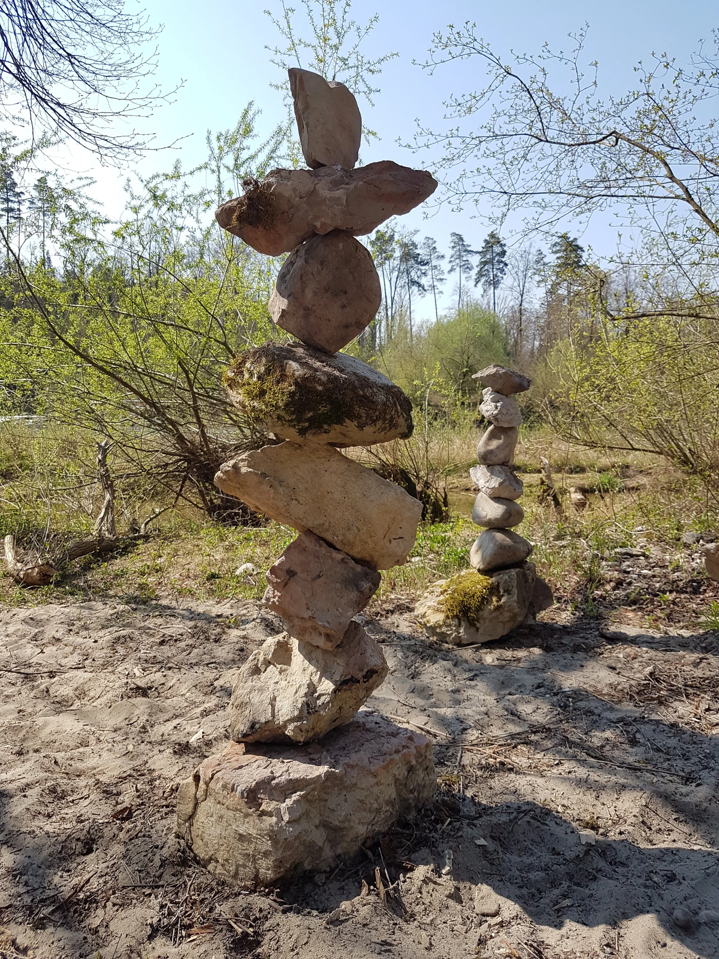 Zwei Steinstapel in einer Naturumgebung mit Bäumen und Sandboden.